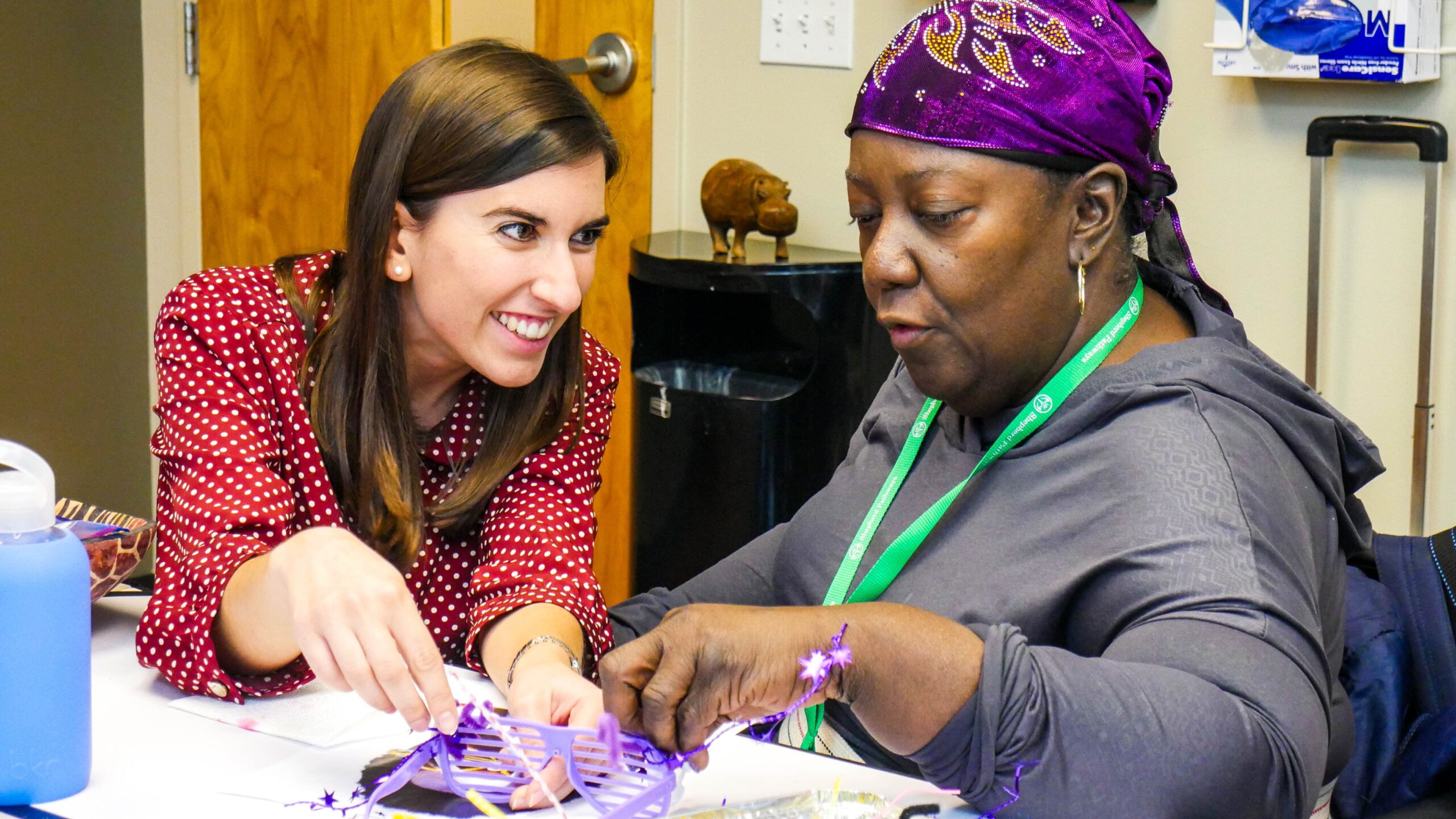 A woman in a red polka dot blouse smiles and assists another woman wearing a purple headscarf. They are engaged in a crafting activity at a table with art supplies.