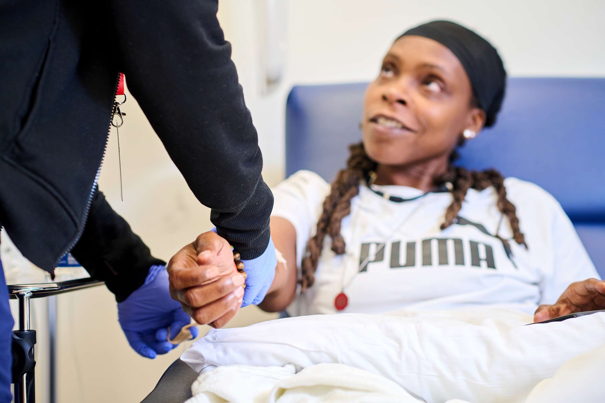 A patient sitting in a chair, looking up and smiling at a gloved healthcare professional who is holding their hand. The patient is wearing a white Puma shirt and a black headband. The healthcare professional is in dark clothing.