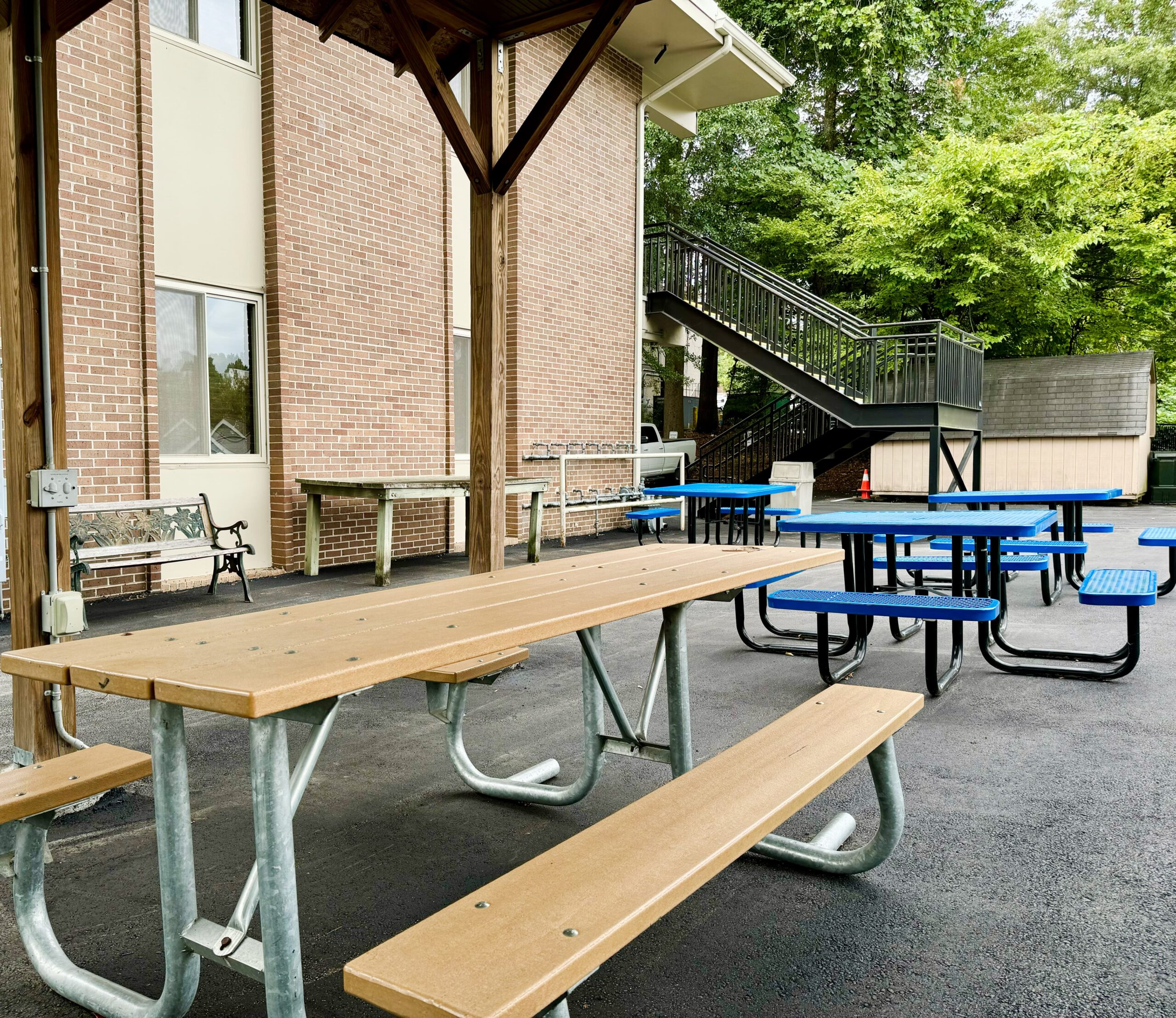 Outdoor area with several picnic tables, some wood and some with blue seats, on a paved surface. Brick building and metal staircase in the background with greenery and a wooden shelter overhead.