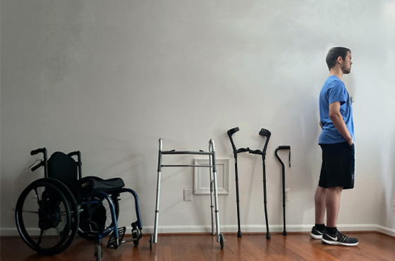 A person stands facing sideways in a room beside a series of mobility aids, including a wheelchair, a walker, crutches, and a cane, indicating a progression of mobility support.