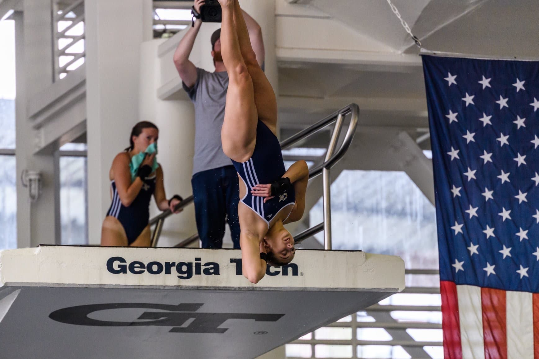 A diver in a blue swimsuit performs a handstand on a diving board labeled 