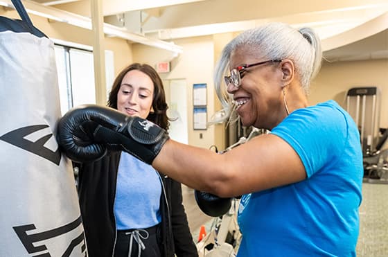 An older woman with gray hair and glasses is smiling and wearing boxing gloves while punching a heavy bag in a gym. A younger woman, also smiling, stands next to her, providing support and guidance. Gym equipment can be seen in the background.