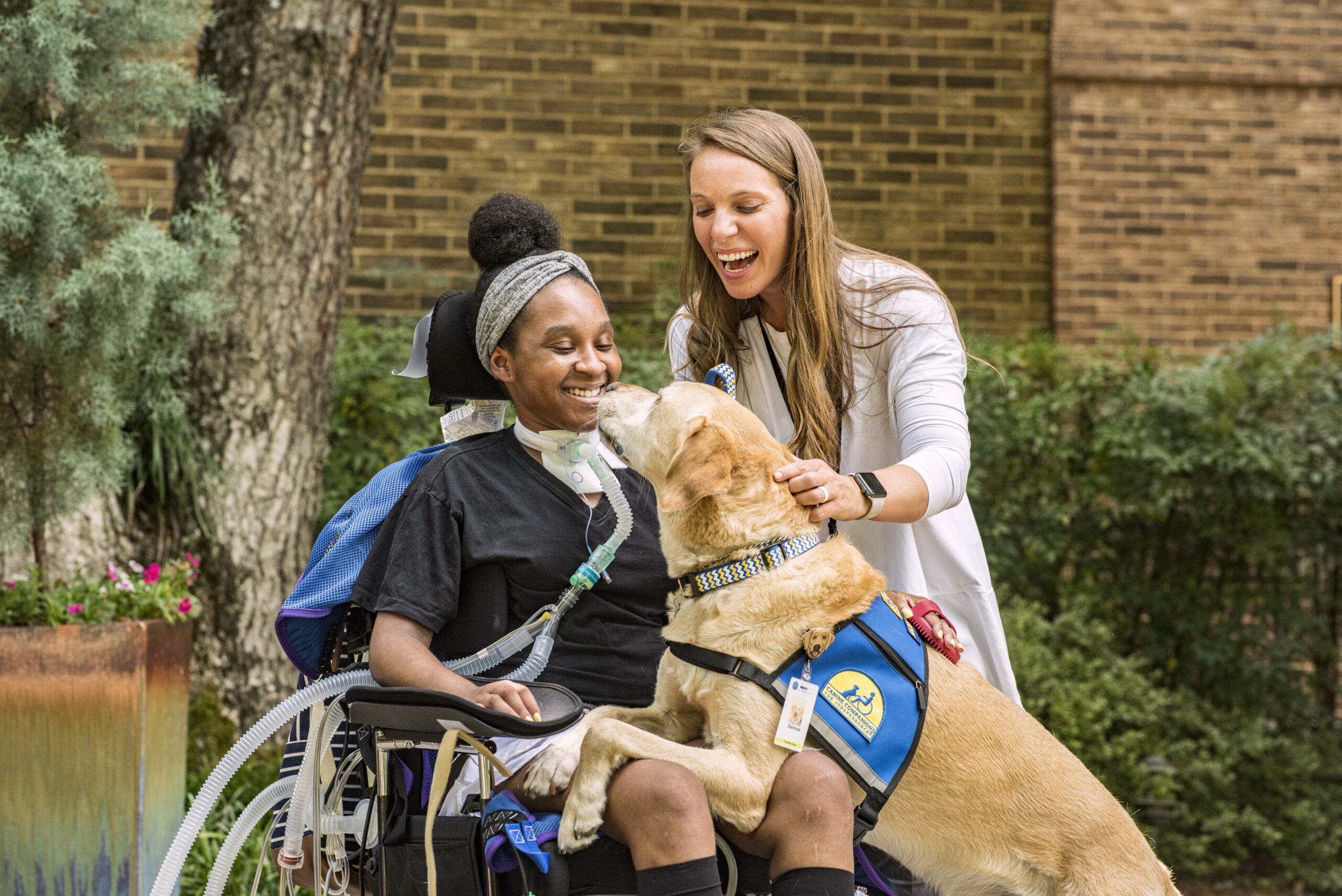 A smiling woman in a wheelchair, wearing black clothes and a headband, happily interacts with a yellow lab service dog that is licking her face. A woman with long hair and a white coat gently touches the dog's back, also smiling. They are outdoors near trees.