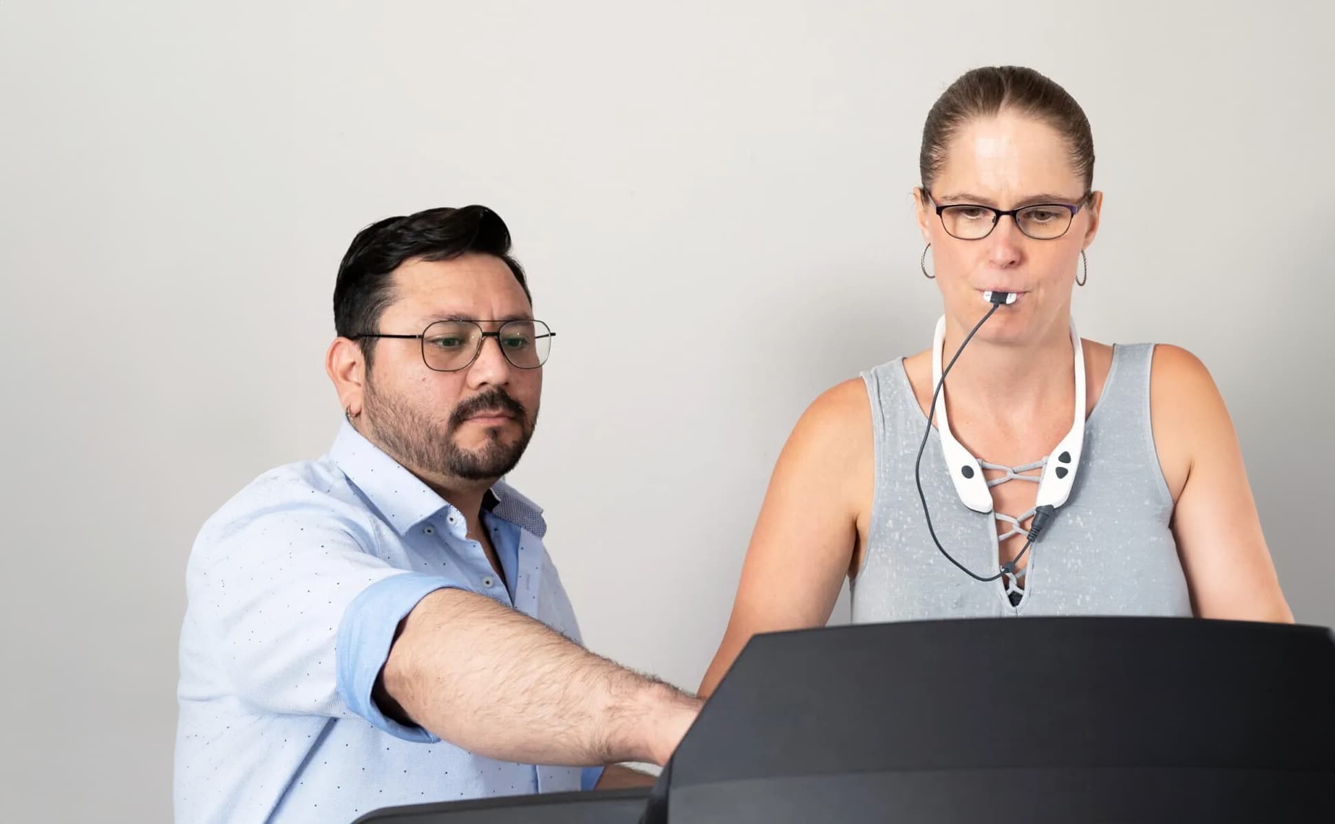 A man in a light blue shirt stands beside a woman in a gray tank top who is wearing a medical device consisting of a neckpiece and a tube attached to her mouth. The man is pointing at something on a screen in front of them, and both are focused on the task.