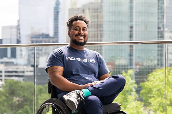 A man with a beard and short curly hair smiles and sits in a wheelchair. He is wearing a blue 