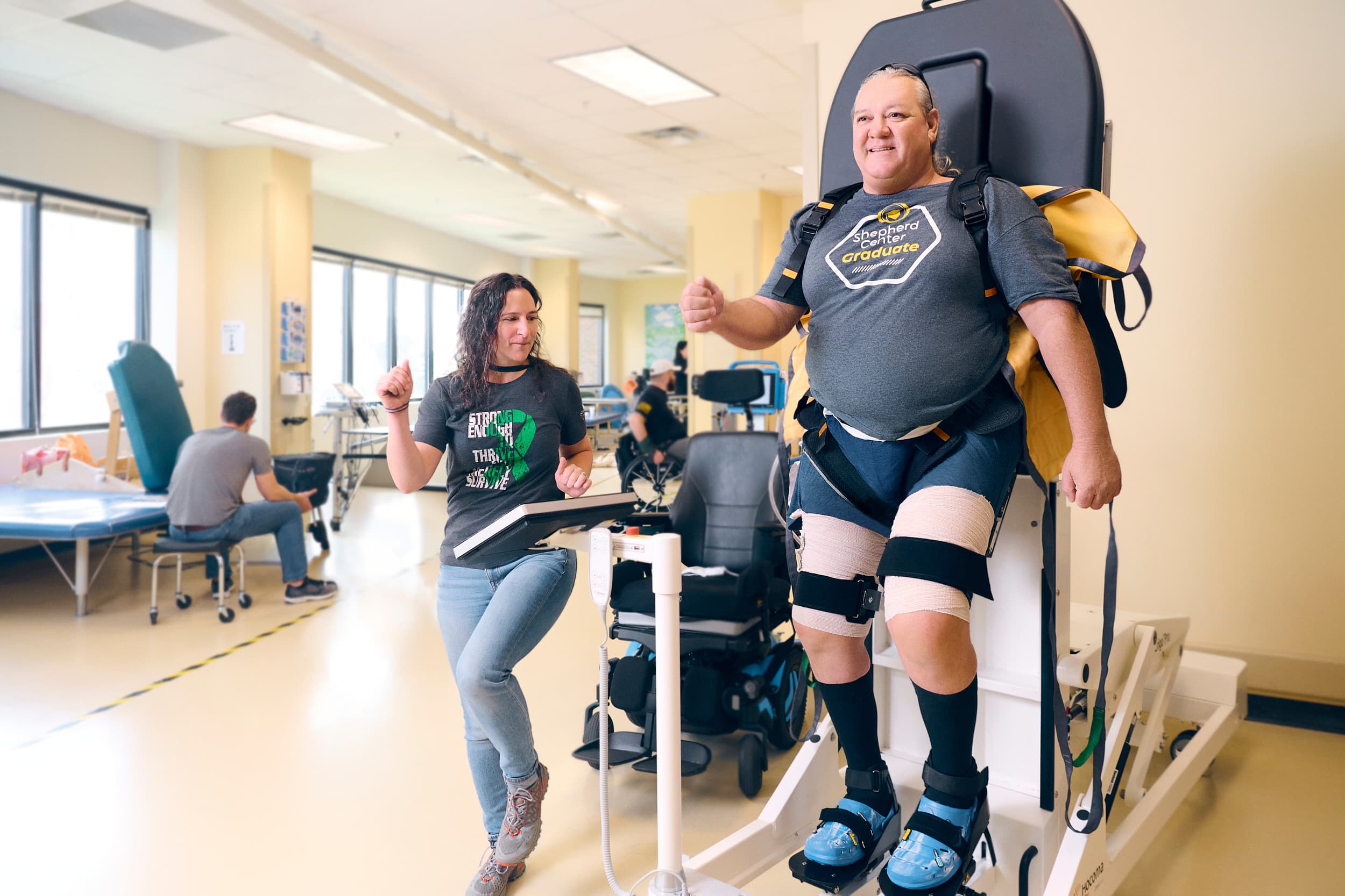 A physical therapist assists a patient using a standing wheelchair in a rehabilitation center. The patient, equipped with supportive straps and knee pads, smiles while the therapist monitors progress on a tablet. Other patients are in the background receiving therapy.