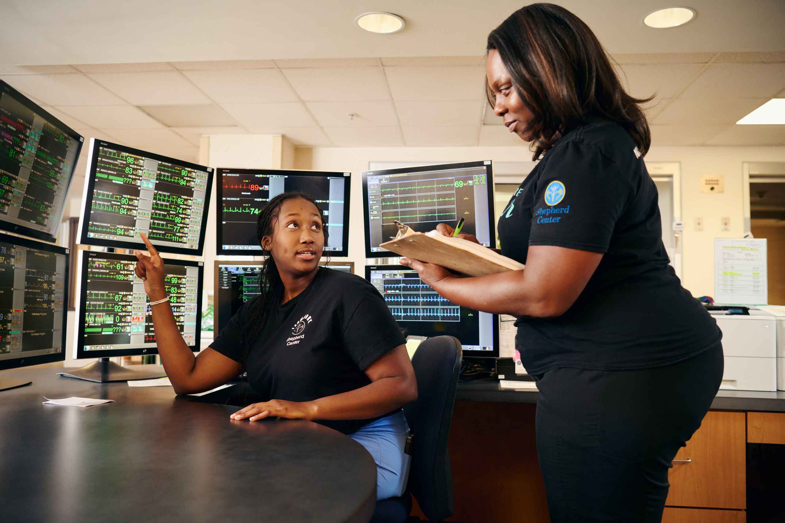 Two women working in a control room with multiple monitors displaying data. One woman is seated, pointing at a screen, while the other stands with a clipboard, engaging in discussion. Both wear black shirts in a professional setting.