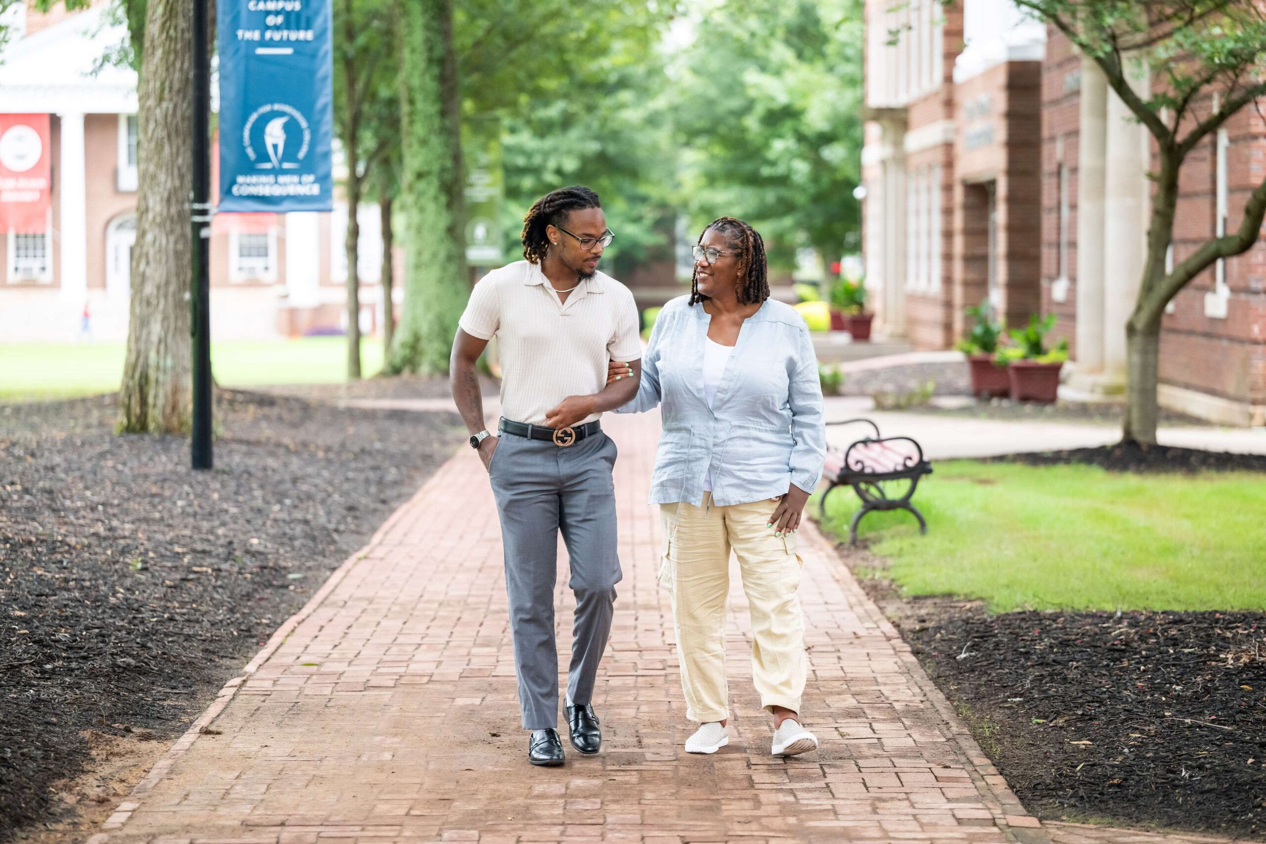 A man and an older woman walk together on a brick pathway in a park-like setting. They are engaged in conversation, with the man looking at the woman. Trees, grass, benches, and buildings are in the background. The man wears a polo shirt and slacks; the woman wears a light blouse and yellow pants.