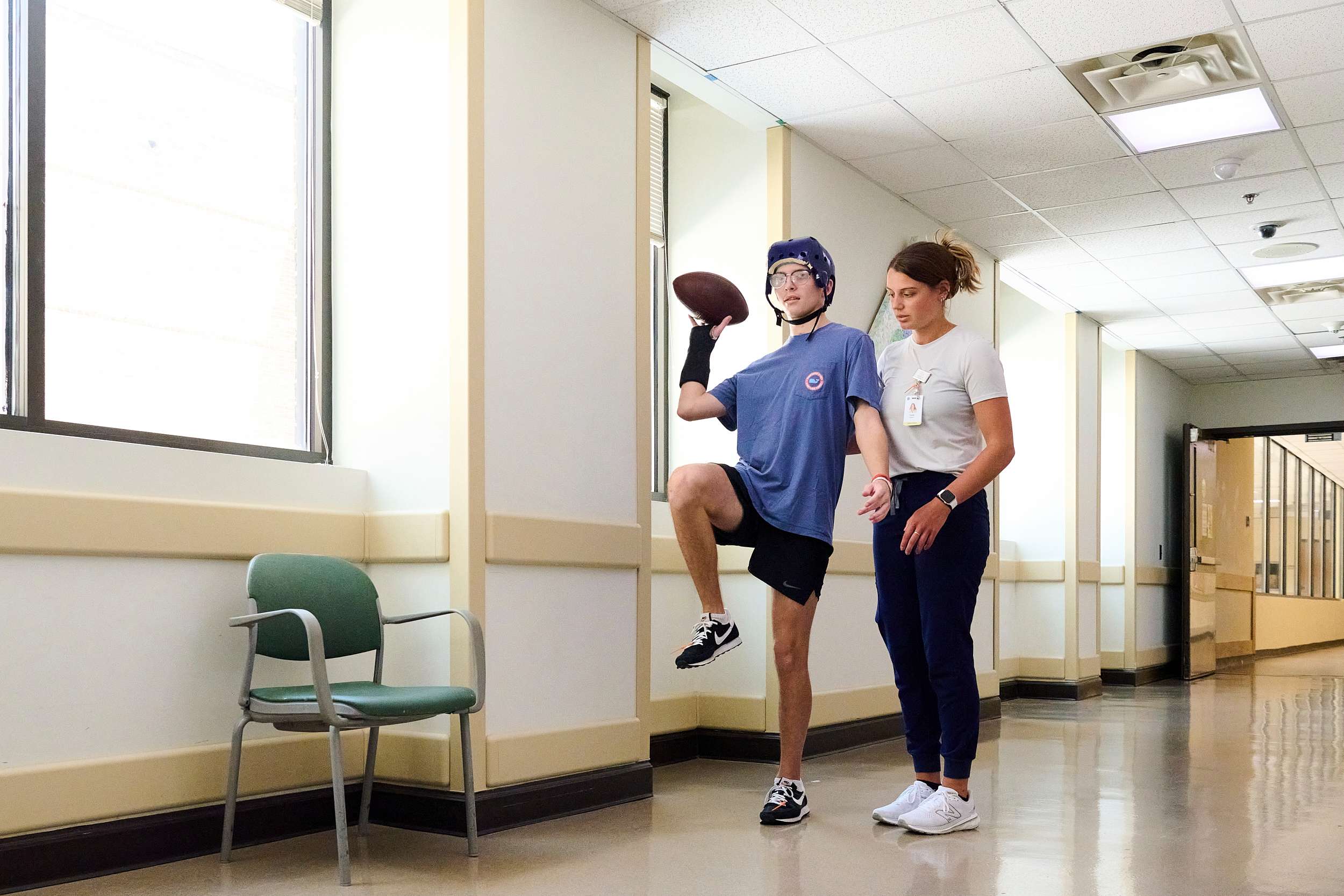 A person wearing a blue helmet and T-shirt practices balance by holding a football and raising one leg, assisted by a therapist. They are in a brightly lit hallway with windows and a green chair nearby.
