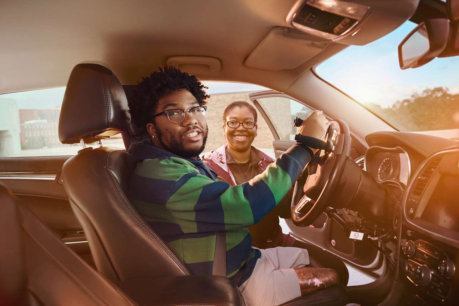 A person with a prosthetic leg sits in the driver's seat of a car, smiling and holding the steering wheel. Another person is in the passenger seat, smiling. Both look towards the camera. Sunlight streams through the window, illuminating the interior.