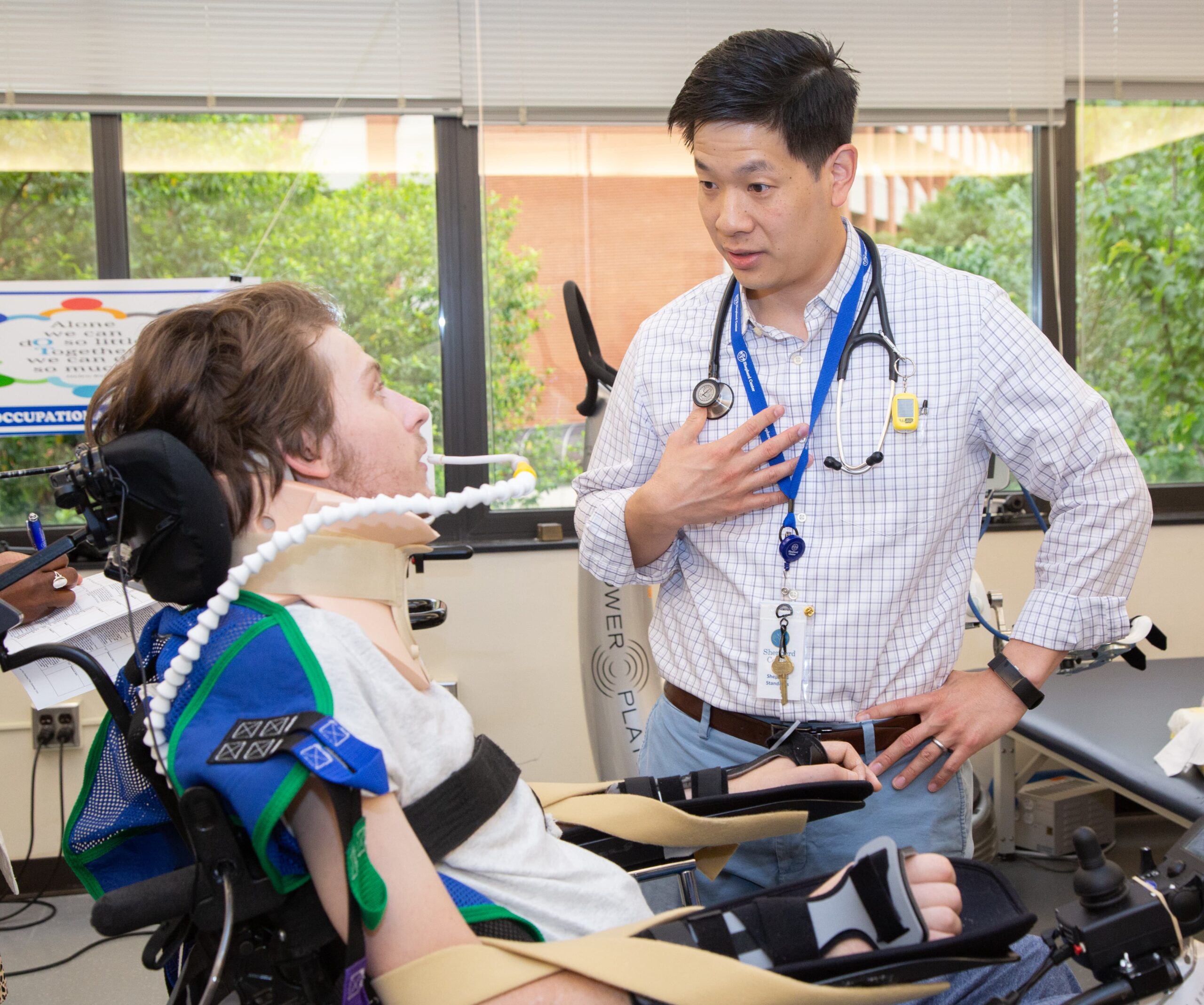 A doctor, wearing a stethoscope and a name badge, talks to a patient in a wheelchair with a breathing tube. The patient is using medical devices and wearing an arm brace. They are in a sunlit room with various equipment and a window view of trees.