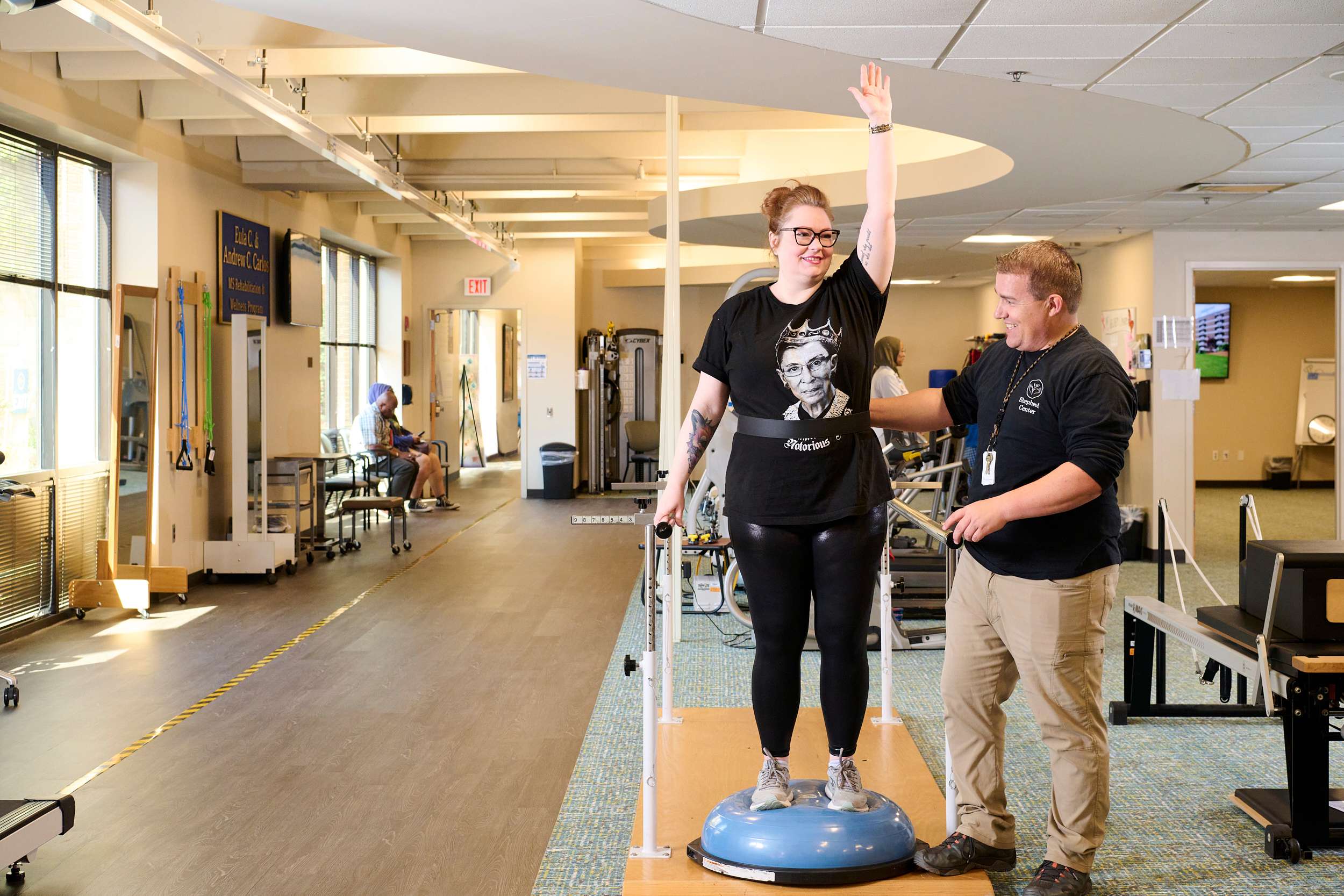 A woman balances on a wobble board in a rehabilitation gym, supported by a man standing beside her. She holds onto a frame with one hand raised. The gym has exercise equipment and a person seated in the background.