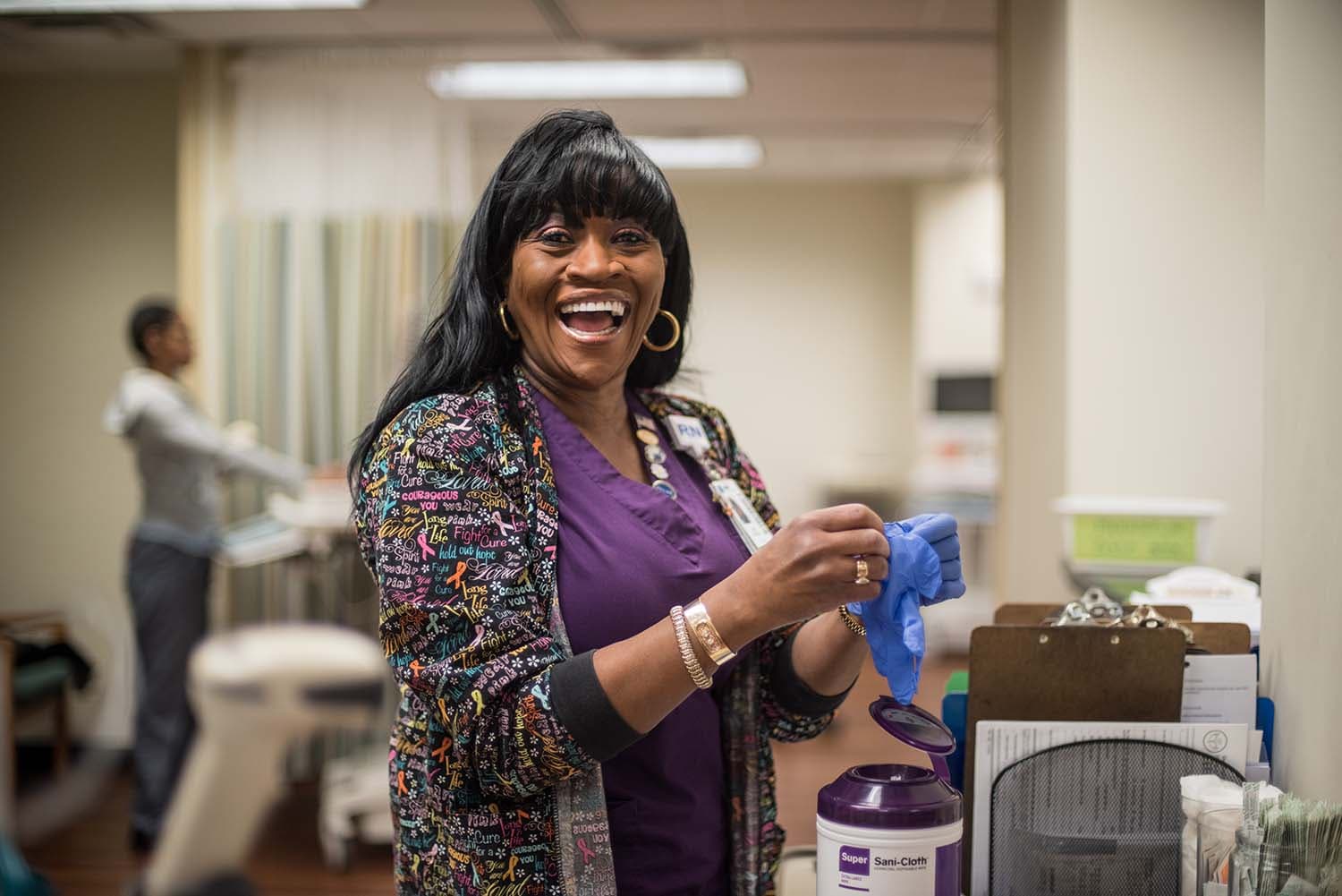 A smiling woman in a colorful cardigan puts on gloves in a medical setting. A person in the background attends to a task. Various medical supplies and equipment are visible around them.