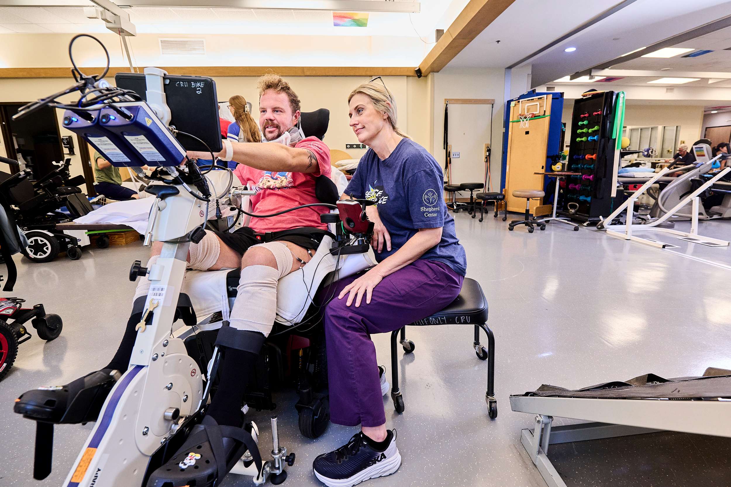 A man with leg braces uses a rehabilitation exercise machine, assisted by a woman in a purple uniform. They are in a physical therapy room with various equipment. Both appear focused on the activity displayed on a screen.