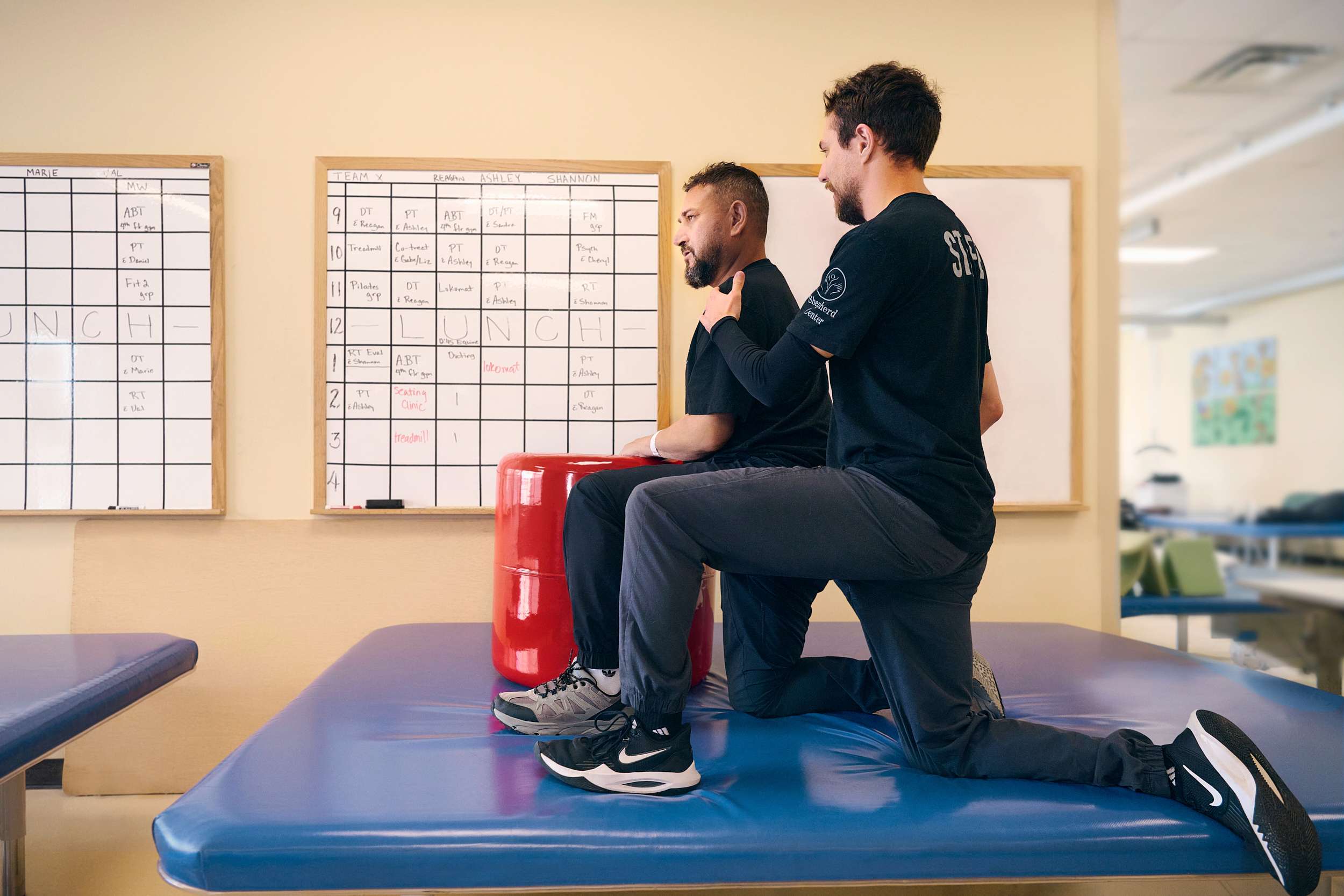 A physical therapist assists a patient with an exercise on a table. The patient sits with back support, while the therapist provides guidance. In the background, whiteboards with schedules are visible. Both wear casual athletic attire.