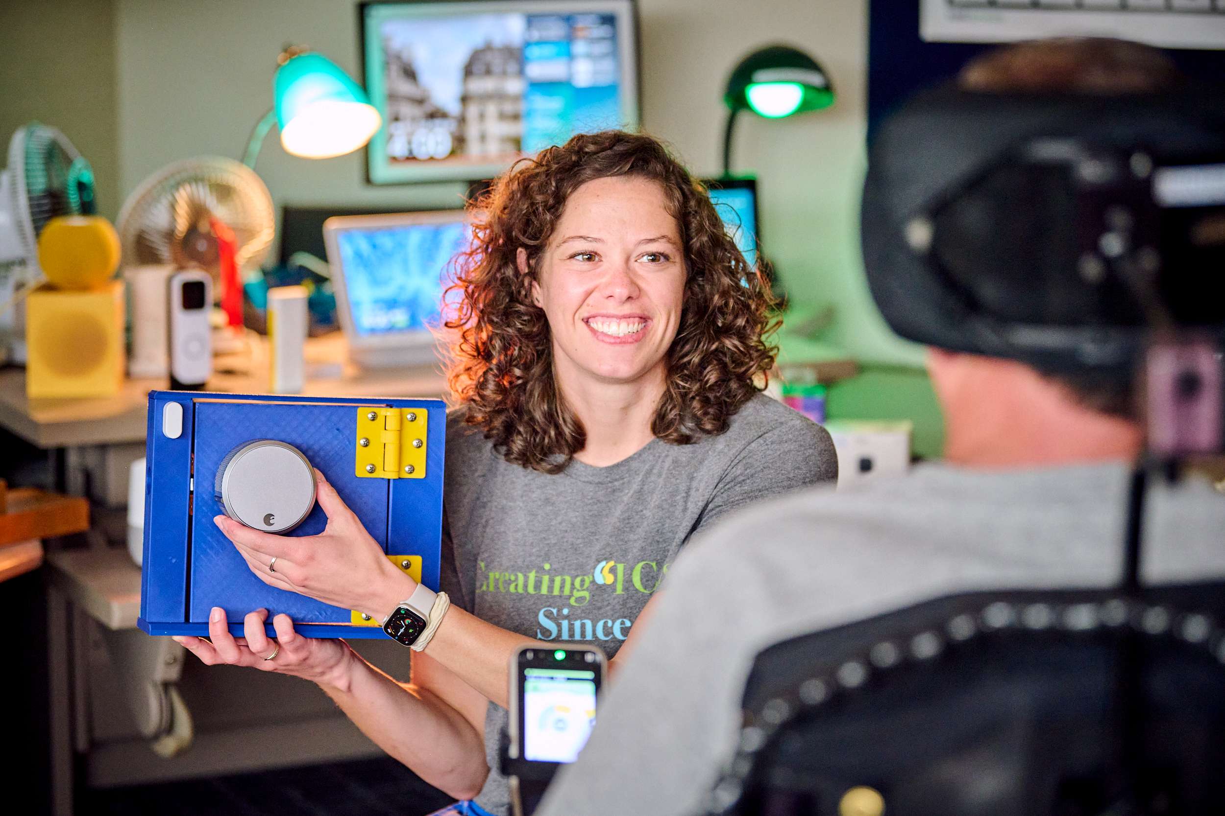 A woman with curly hair smiles at a person in a wheelchair. She is holding a colorful electronic device with dials. The setting is a room filled with computers and tech equipment, creating a friendly and tech-focused atmosphere.