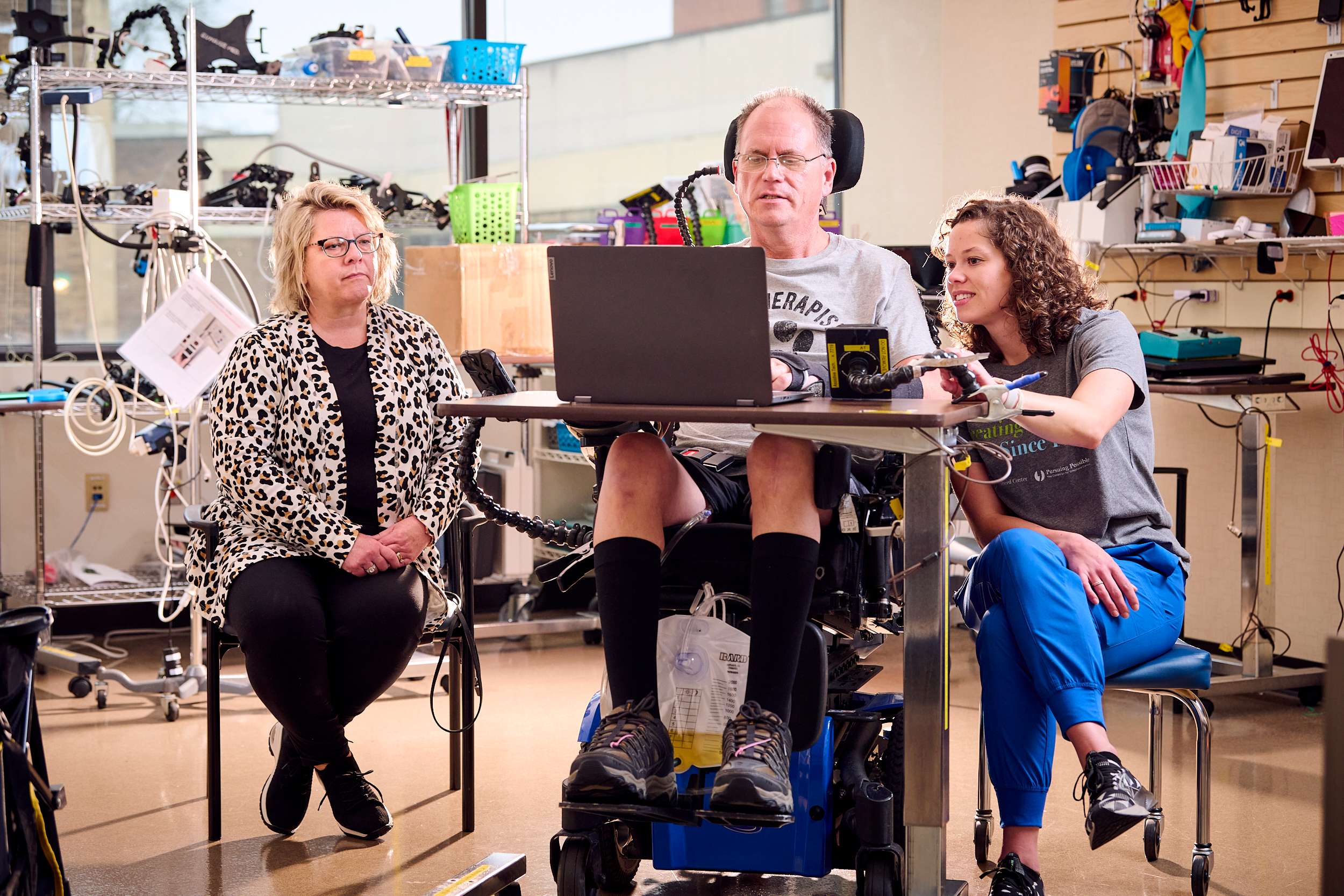 A man in a wheelchair uses technology at a table, assisted by a woman sitting nearby. Another woman sits to the left. The room is equipped with various tech devices and tools, suggesting a workshop or lab environment.