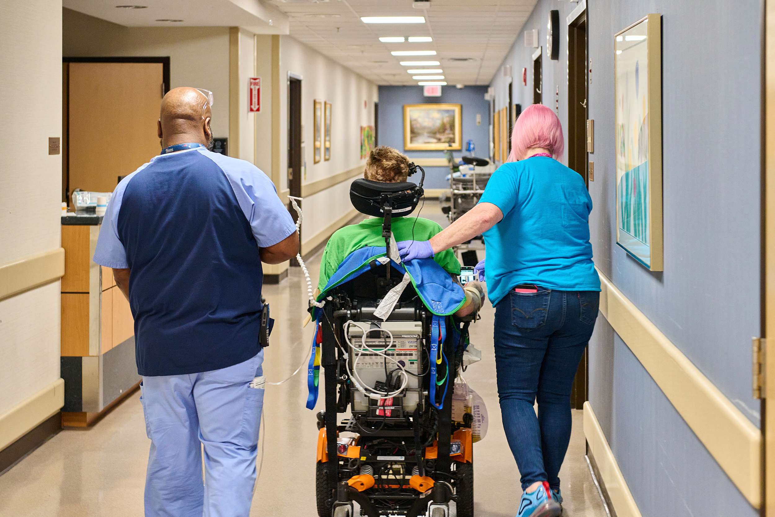 Two respiratory therapists assist a patient in a motorized wheelchair down a hospital corridor. One provides support from behind, and the other walks alongside.