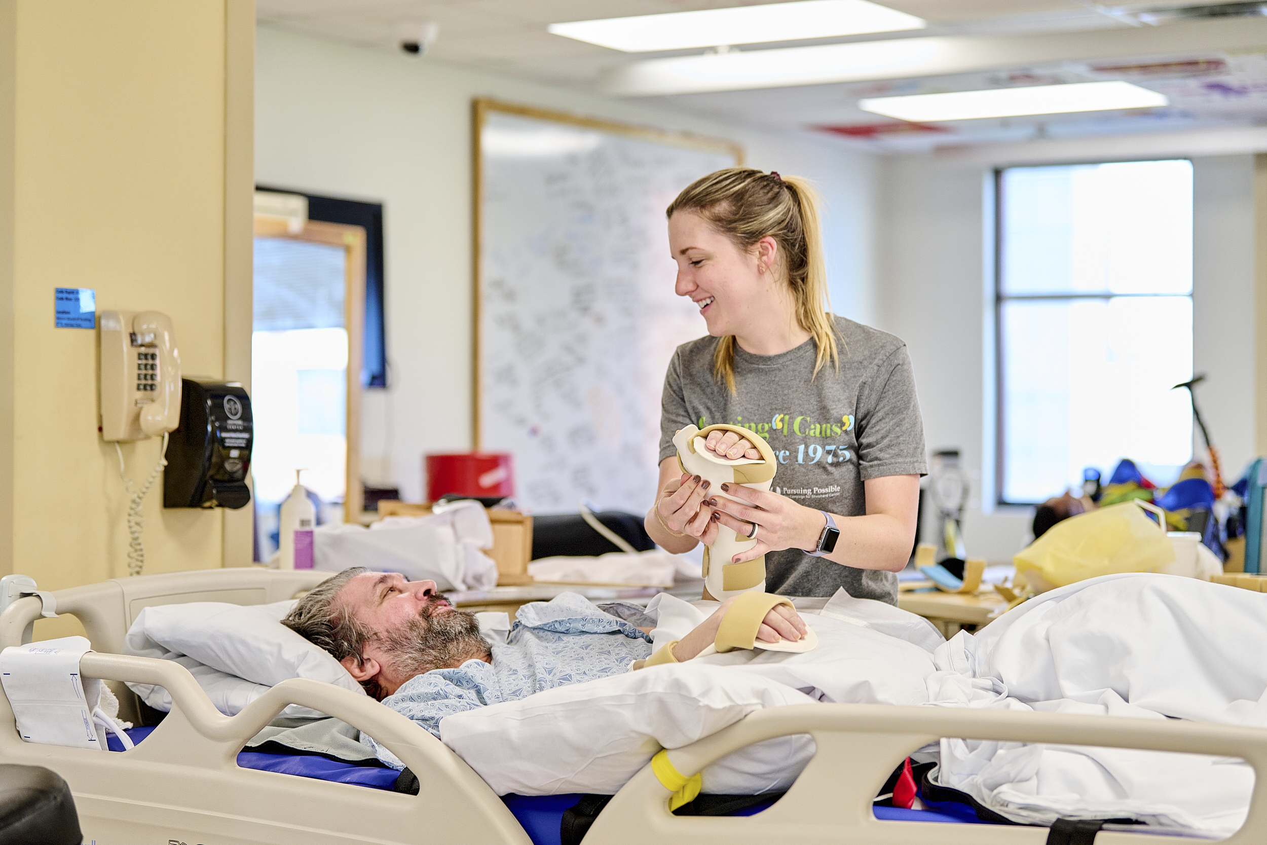 A nurse assists a smiling patient in a hospital bed with hand exercises using a foam roller. The room has medical equipment, a wall phone, and a large window letting in natural light.