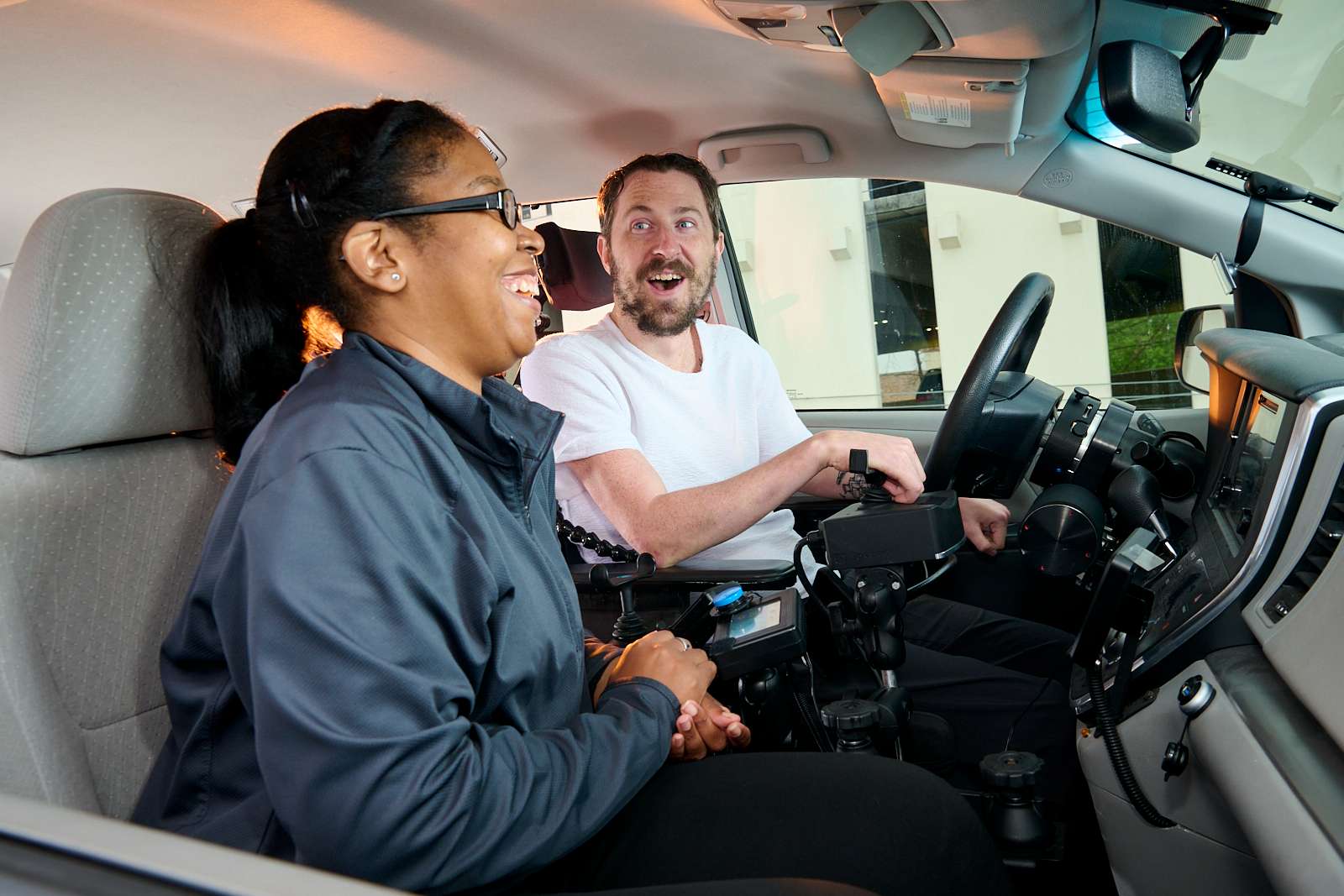 A man in a wheelchair-accessible van is smiling and engaging with a woman sitting in the passenger seat. The woman is wearing glasses and a jacket, and both appear happy and engaged in conversation.