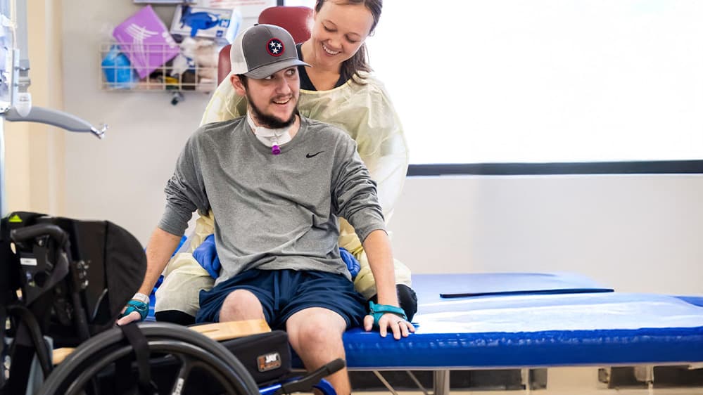 A person wearing a gray cap and casual sportswear is smiling while sitting on a therapy table. A caregiver in a protective gown stands behind, assisting. A wheelchair is nearby, and medical equipment is visible in the background.
