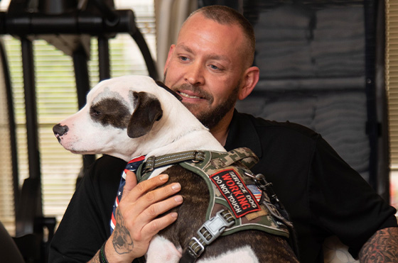 A man sits smiling, holding a white and brown dog wearing a green vest with a 