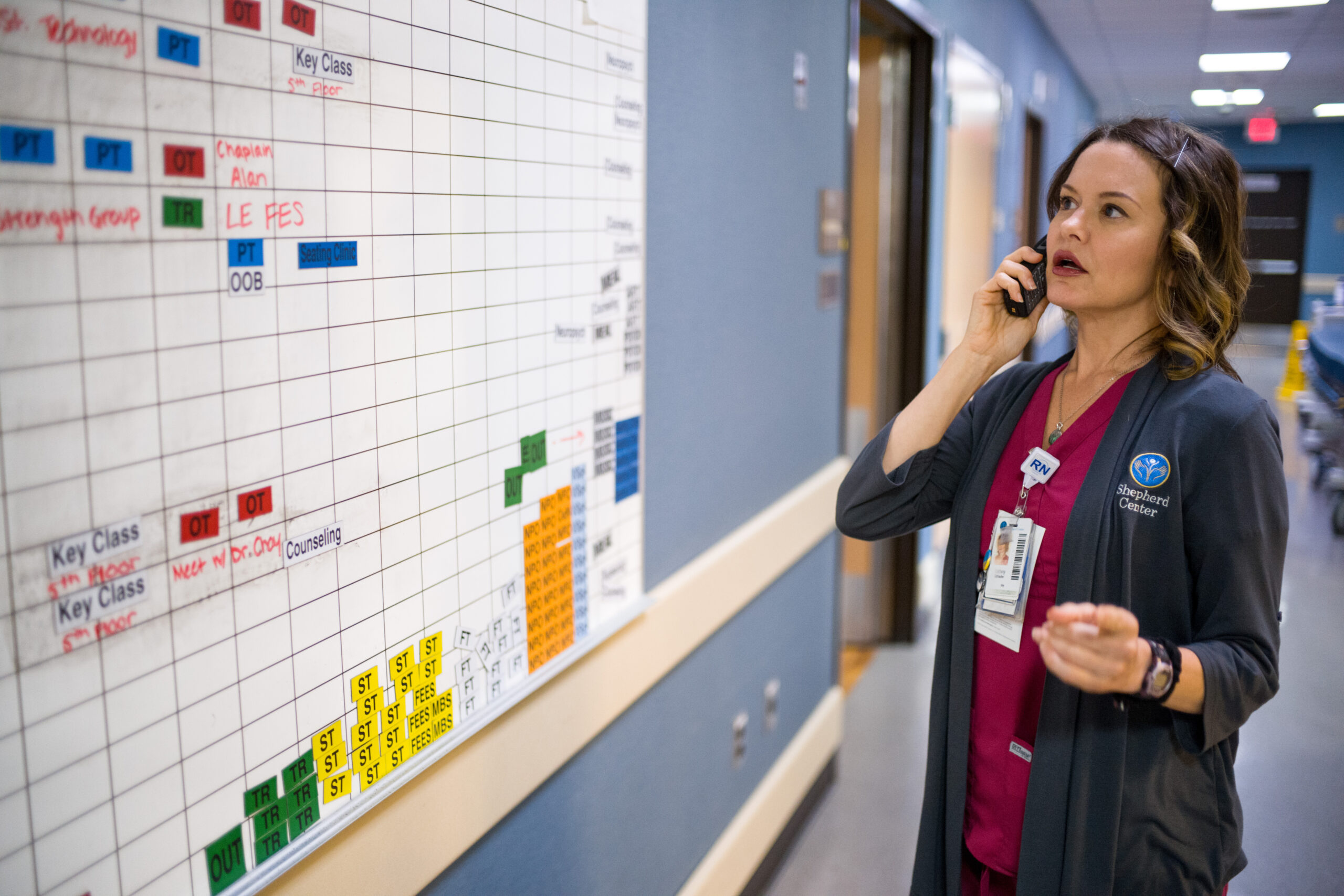 A healthcare worker in scrubs and a coat is talking on the phone while pointing at a large scheduling board in a hospital hallway. The board is covered with color-coded notes and columns.