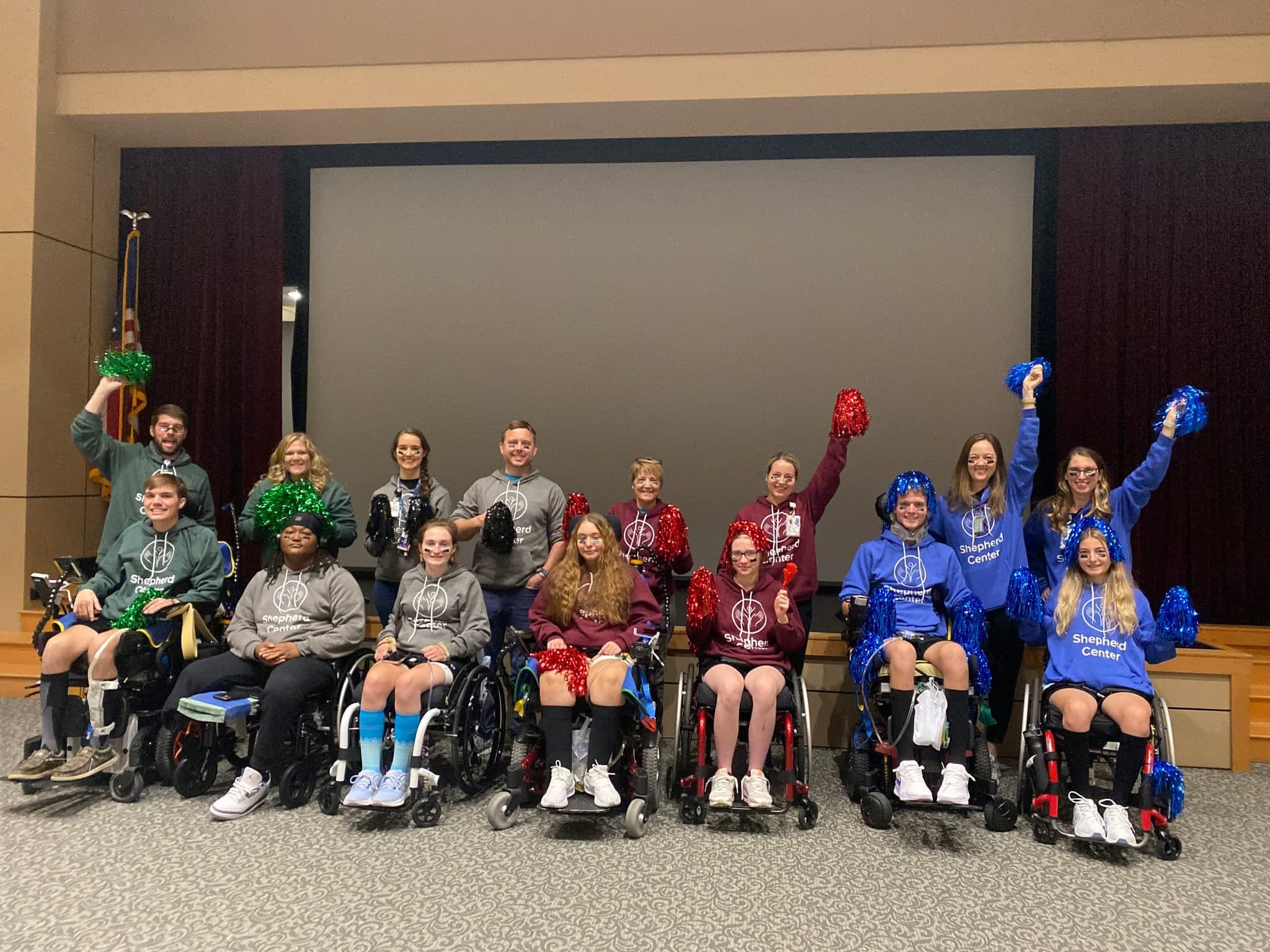 A group of people in wheelchairs smiling and holding colorful pom-poms. They are in a room with a large screen behind them. Some wear sweatshirts with logos, and they appear to be celebrating or participating in an event.