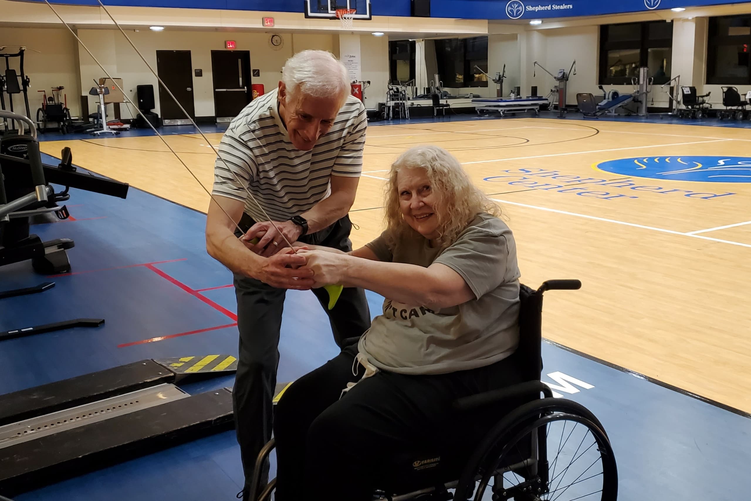 A man helps a woman in a wheelchair aim a bow in an indoor sports facility. The gym has a basketball court and exercise equipment. The woman is smiling, and both appear focused on the activity.