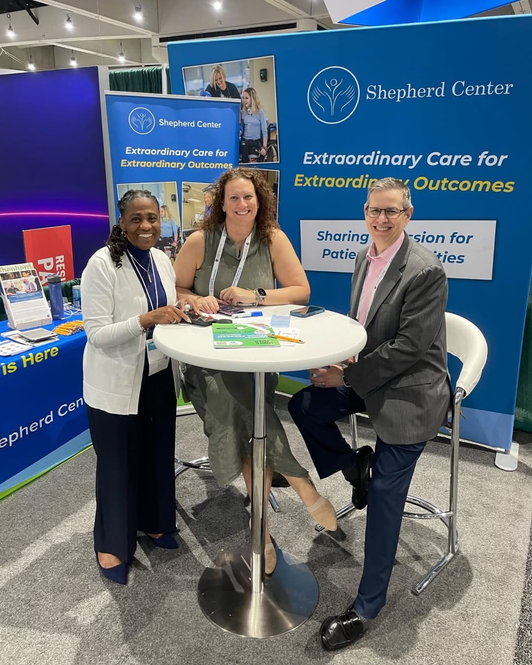 Three people smiling at a promotional booth for Shepherd Center. Two women and a man are standing and seated around a high table, with brochures and a display reading 