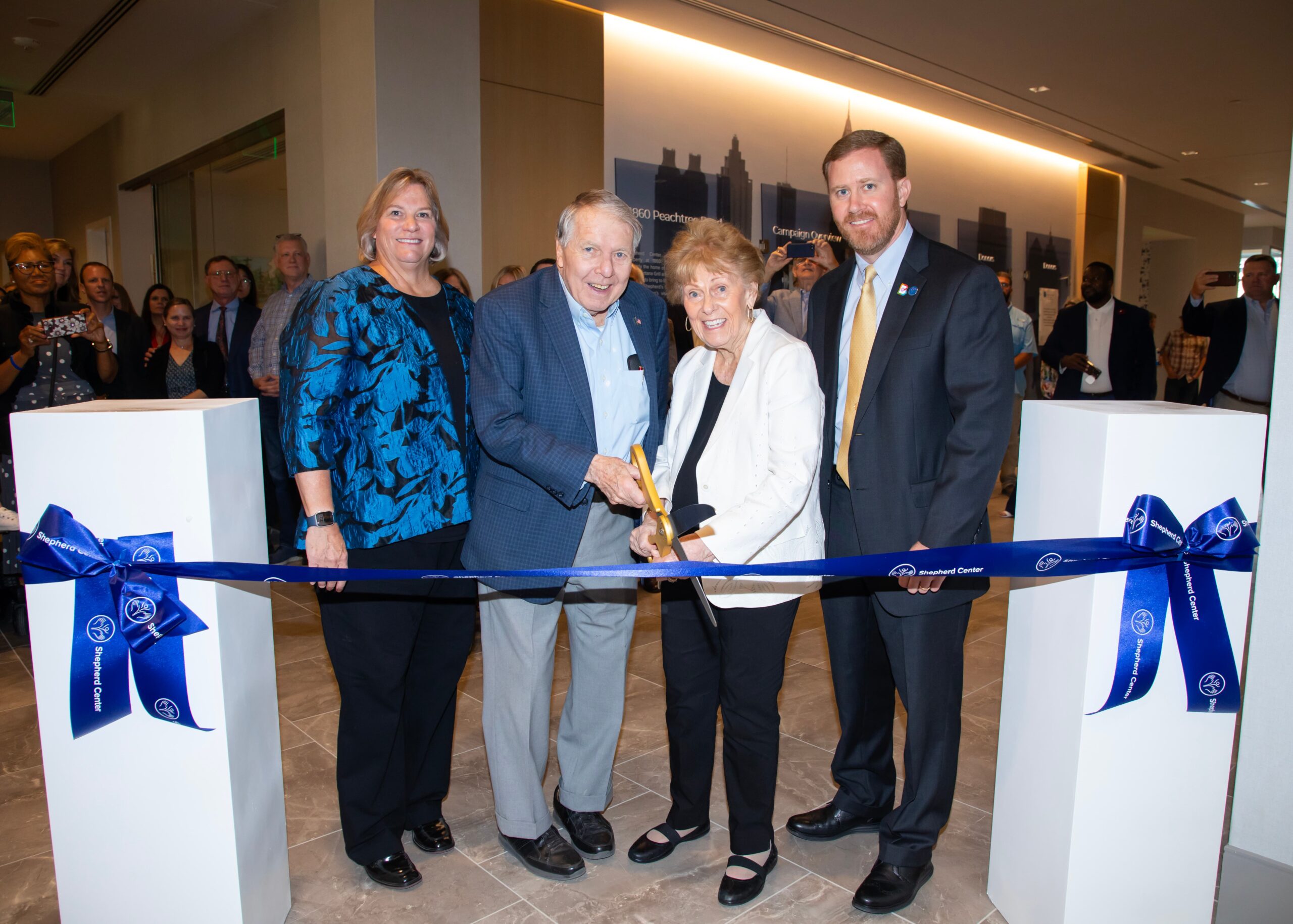 Four people smile while holding large scissors to cut a blue ribbon in an indoor event. A crowd is visible in the background, and the setting appears to be a modern building or office space. The event seems to be a ribbon-cutting ceremony.