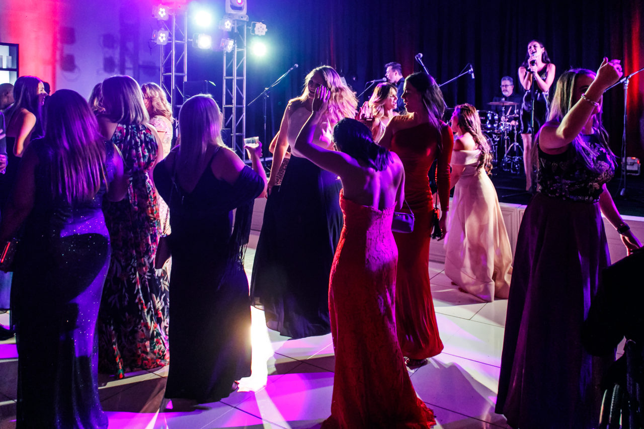 A lively dance floor scene with people in formal dresses dancing. A band performs in the background, illuminated by colorful stage lights that add vibrancy to the atmosphere.