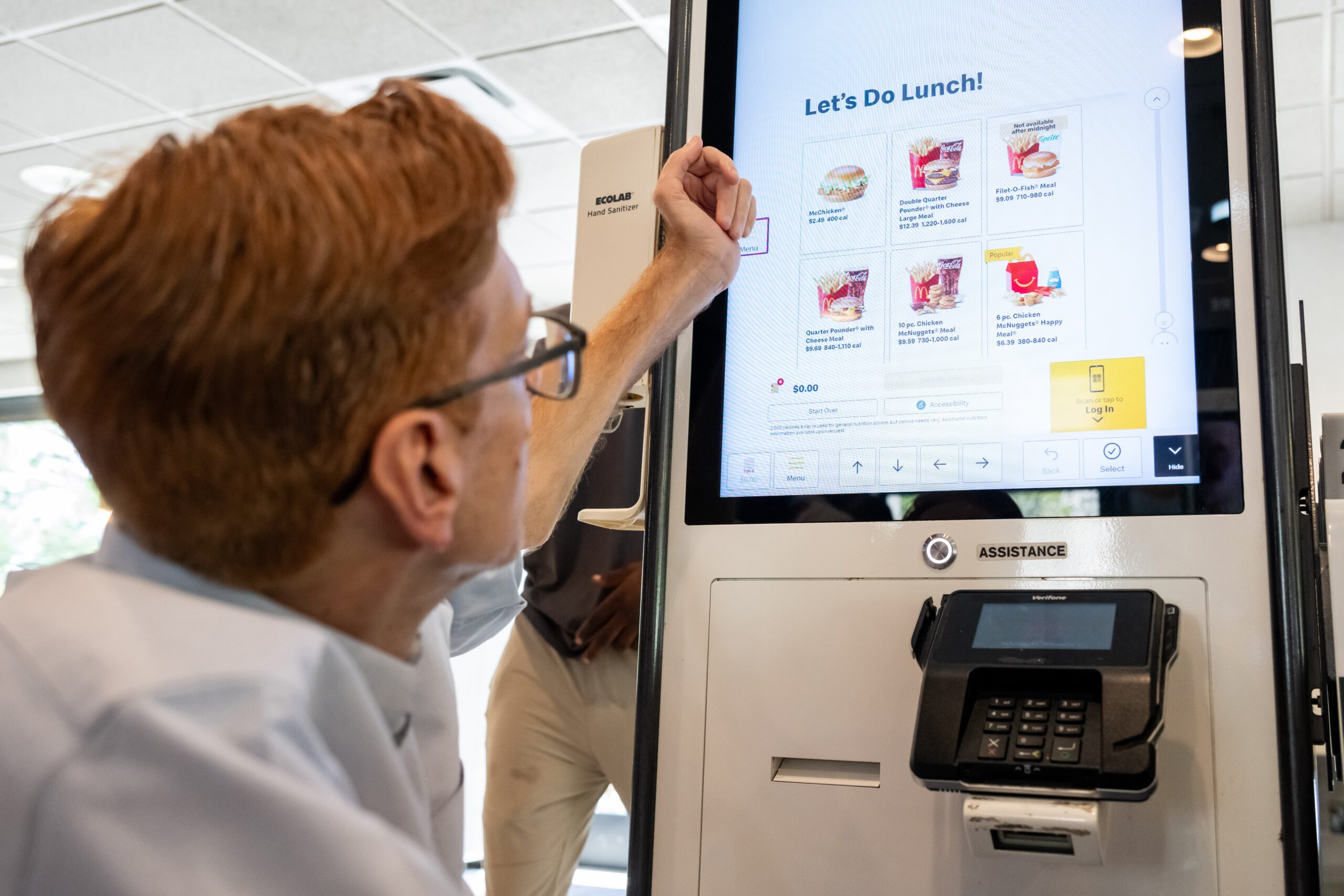 A person with red hair uses a touchscreen ordering kiosk at a restaurant, selecting lunch menu items displayed on the large screen.