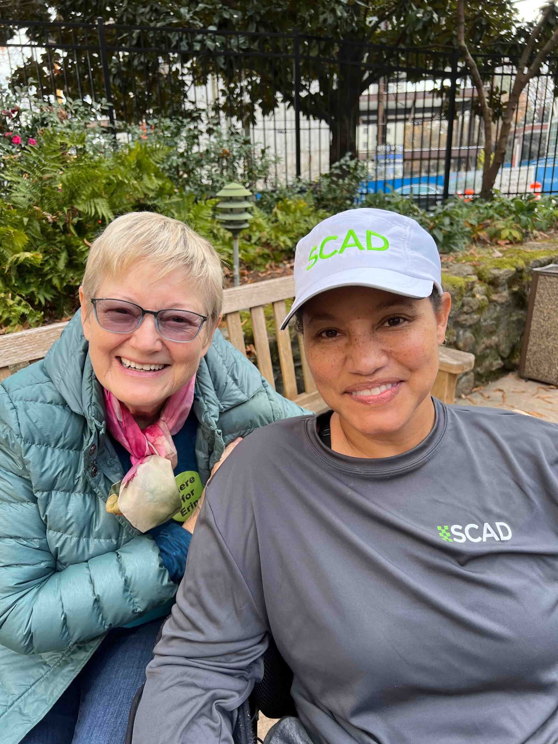 Two people are smiling outdoors. One is wearing a green jacket and pink scarf, while the other wears a gray shirt and cap with 