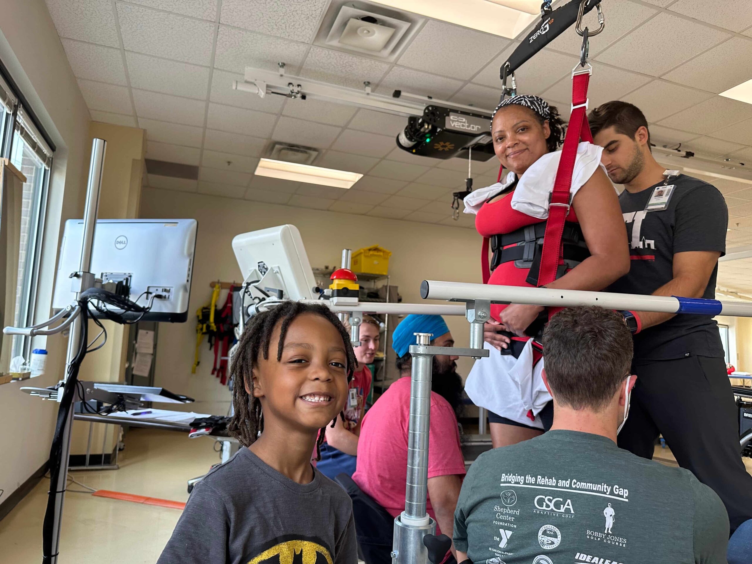 A woman is suspended in a harness inside a physical therapy setting. She is smiling, with a man assisting her. A child in the foreground smiles at the camera. Equipment and a computer are visible in the room.