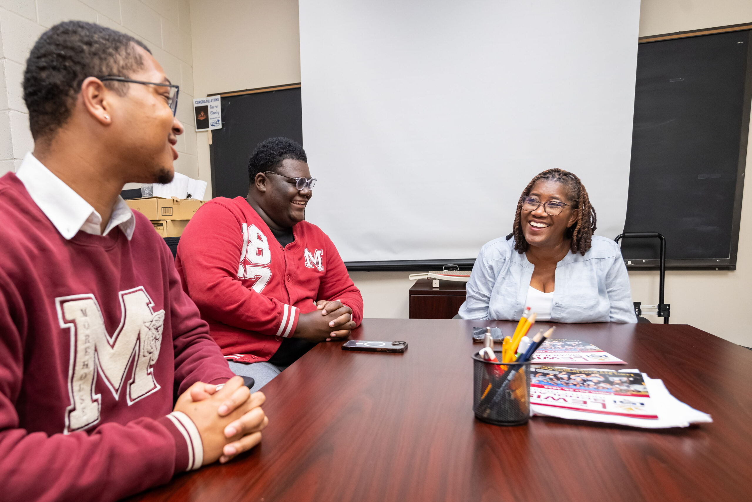Three people sit at a table in a classroom setting, engaged in conversation and smiling. Two wear red letterman jackets with a white 