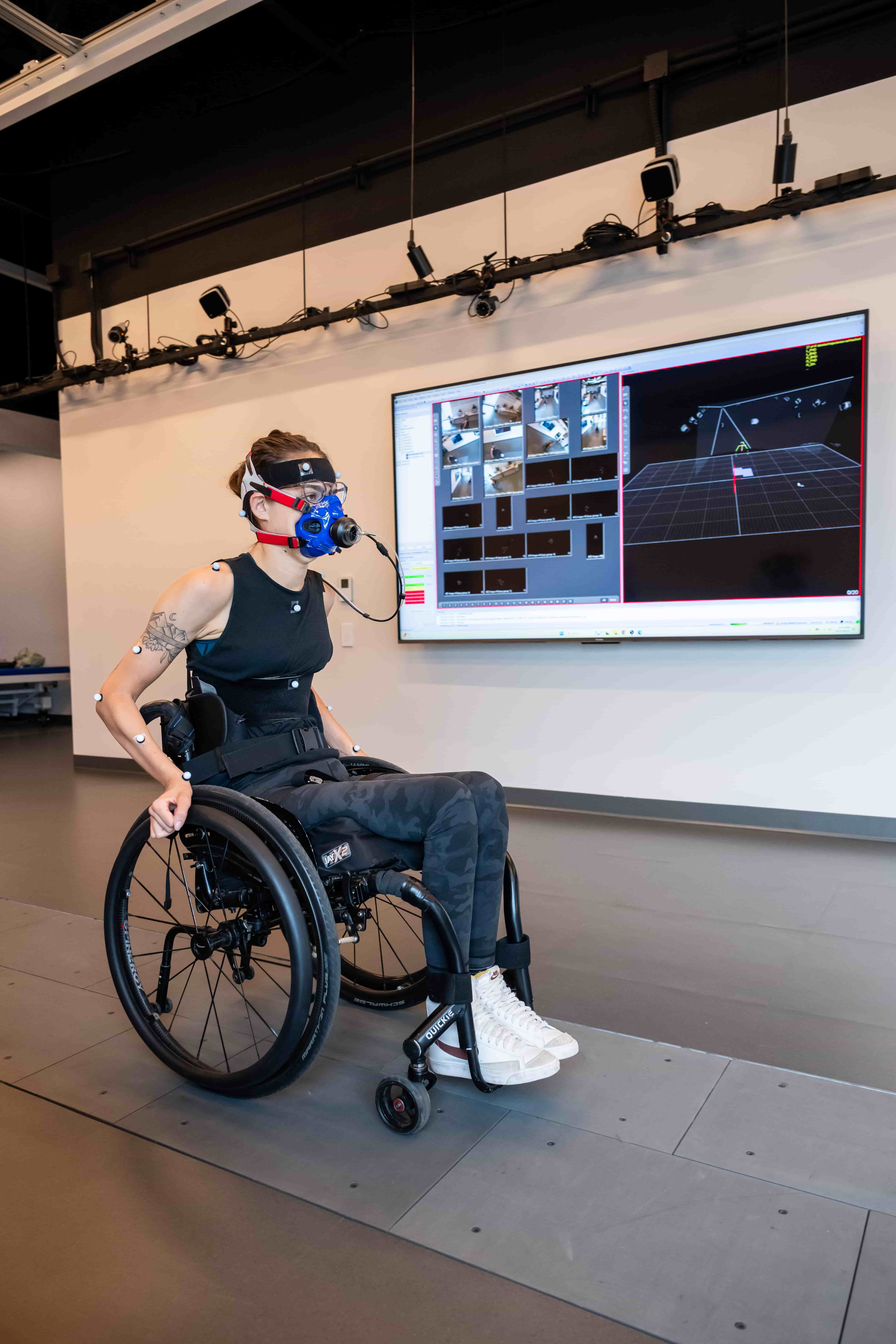 A person in a wheelchair wears a mask with sensors and electrodes, moving along a track in a lab. A large screen behind displays motion capture and data analysis visuals.