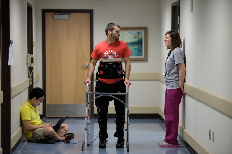 A man using a walker and wearing a robotic exoskeleton stands in a hallway, talking to a woman. Another person sits on the floor nearby, working on a laptop. The scene appears to be in a medical or rehabilitation facility.
