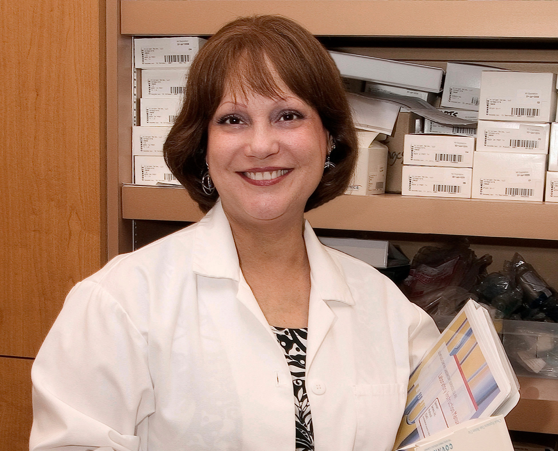 A woman in a white lab coat smiles while holding folders, standing in front of shelves filled with boxes and medical supplies.
