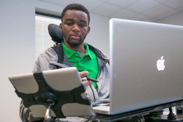 A man using a wheelchair works at a desk with a MacBook and a tablet. He holds a stylus and looks at the tablet, appearing focused. The tablet is mounted on his wheelchair, and there is a window in the background.