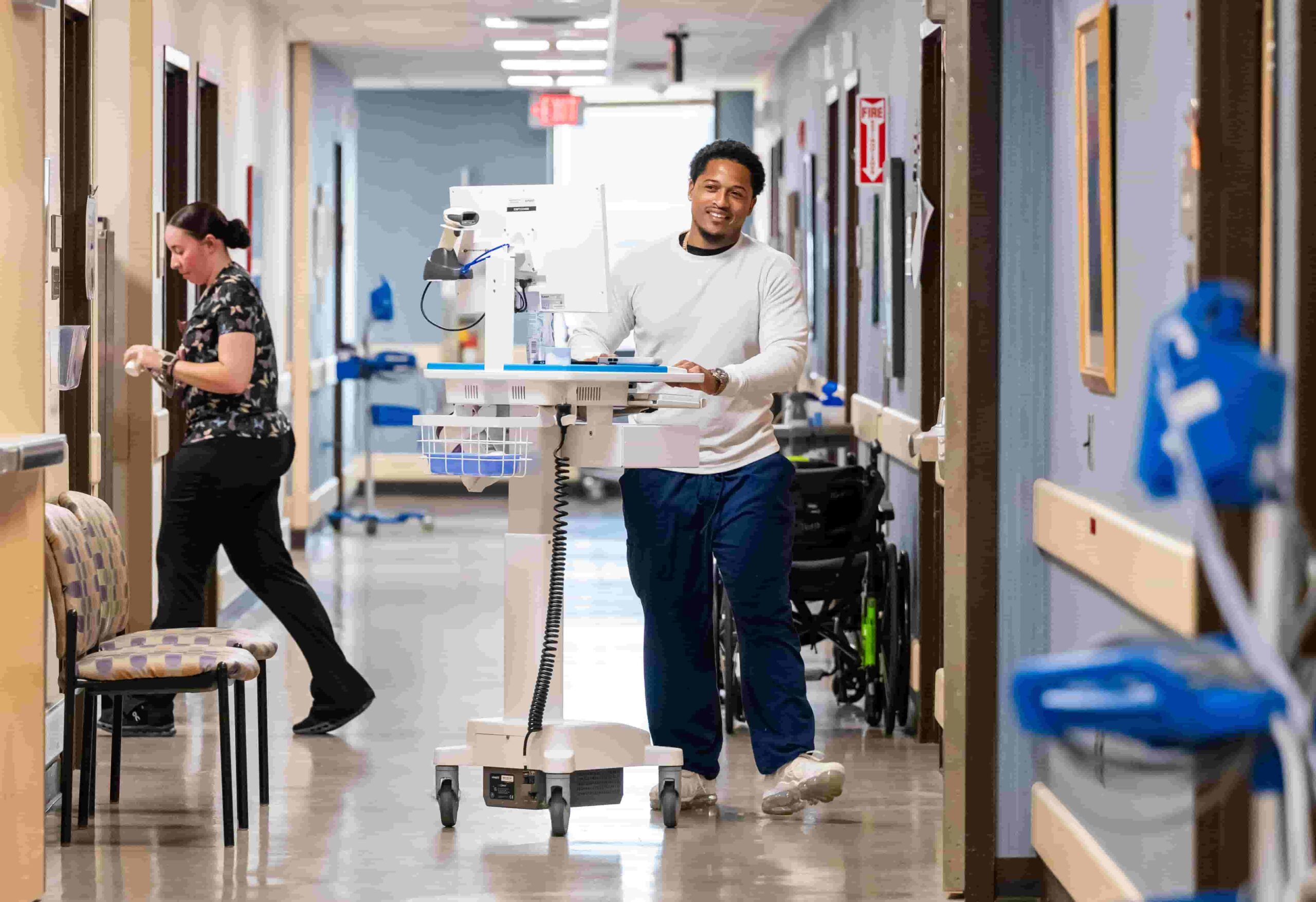 A healthcare worker pushes a mobile workstation down a brightly lit hospital hallway, while another person walks away in the background, both engaged in their tasks.