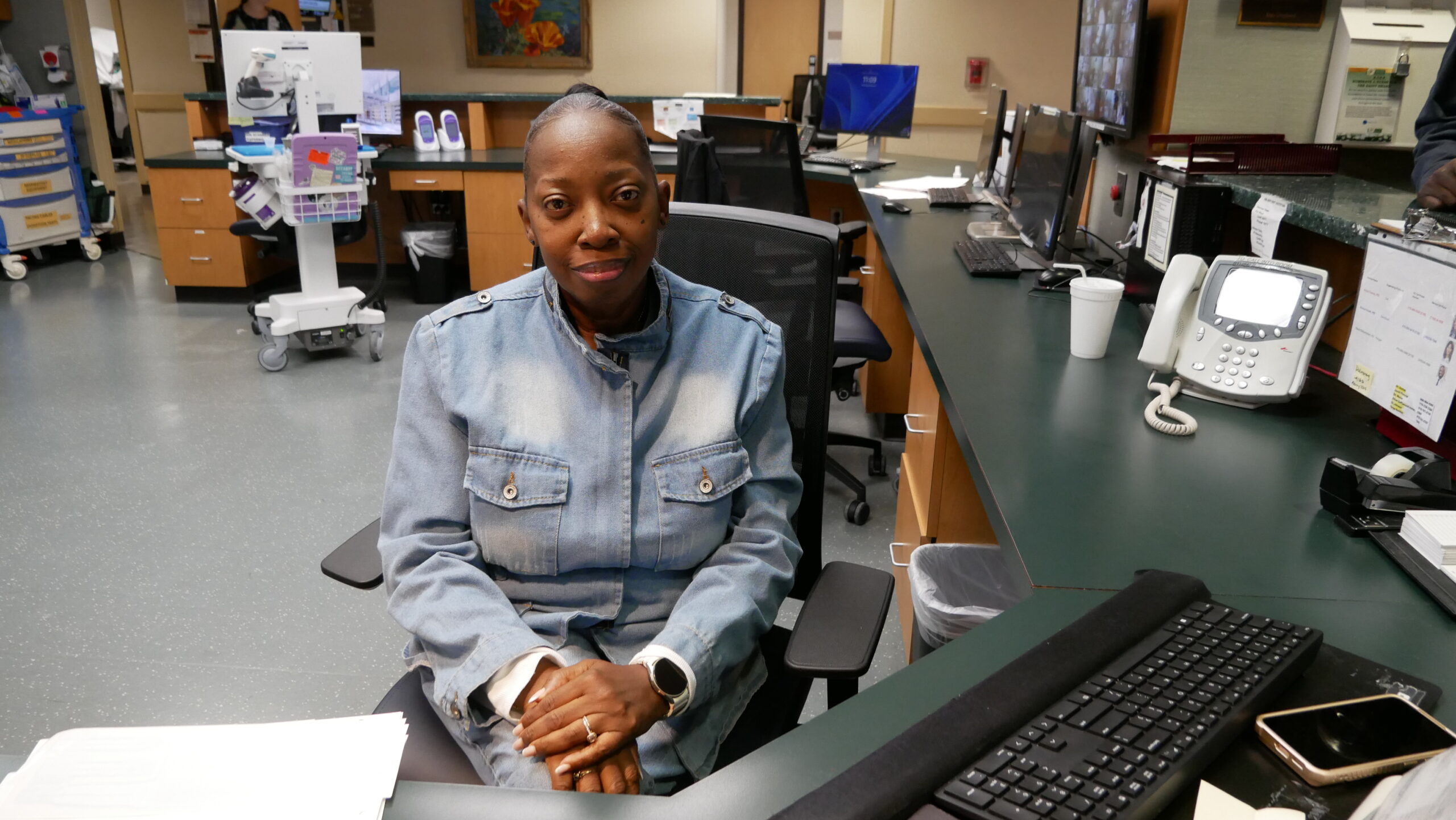 Matilda Lamar-Reed wearing a denim jacket sits at a desk surrounded by computers, phones, and office equipment. She faces the camera with her hands folded on her lap.