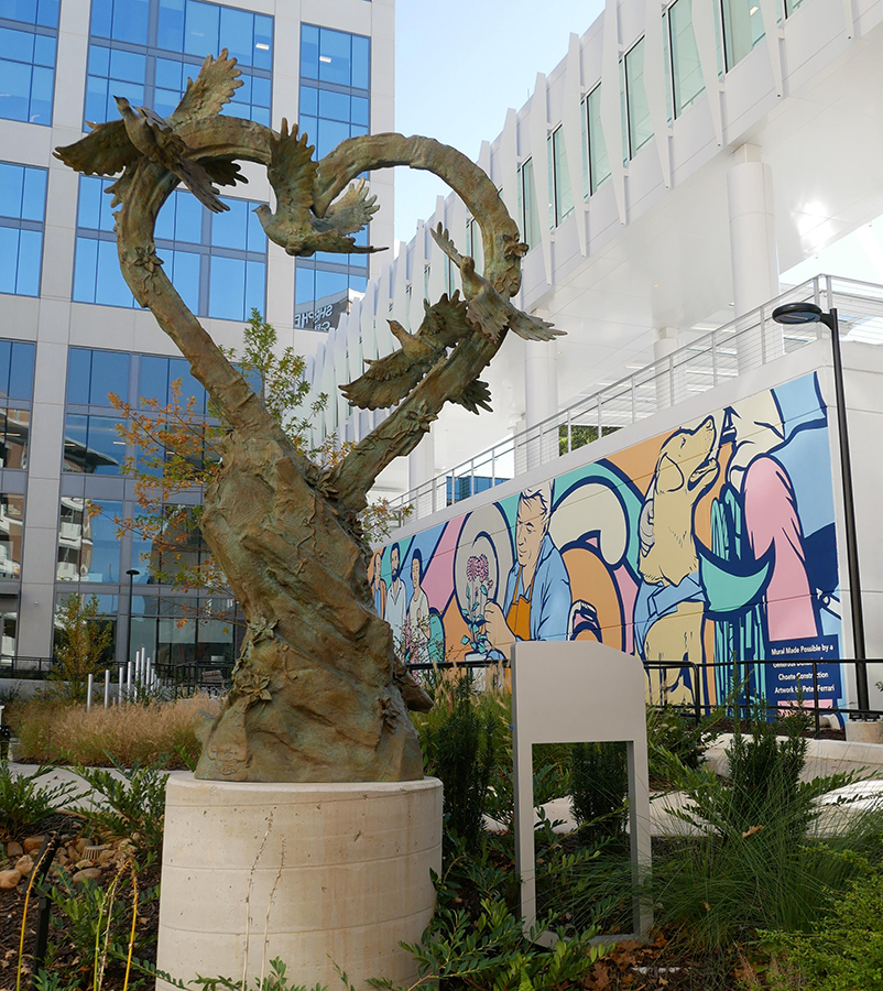 A bronze sculpture shaped like a heart with birds sits in front of a modern building and a colorful mural depicting people and dogs. Grassy landscaping surrounds the sculpture.