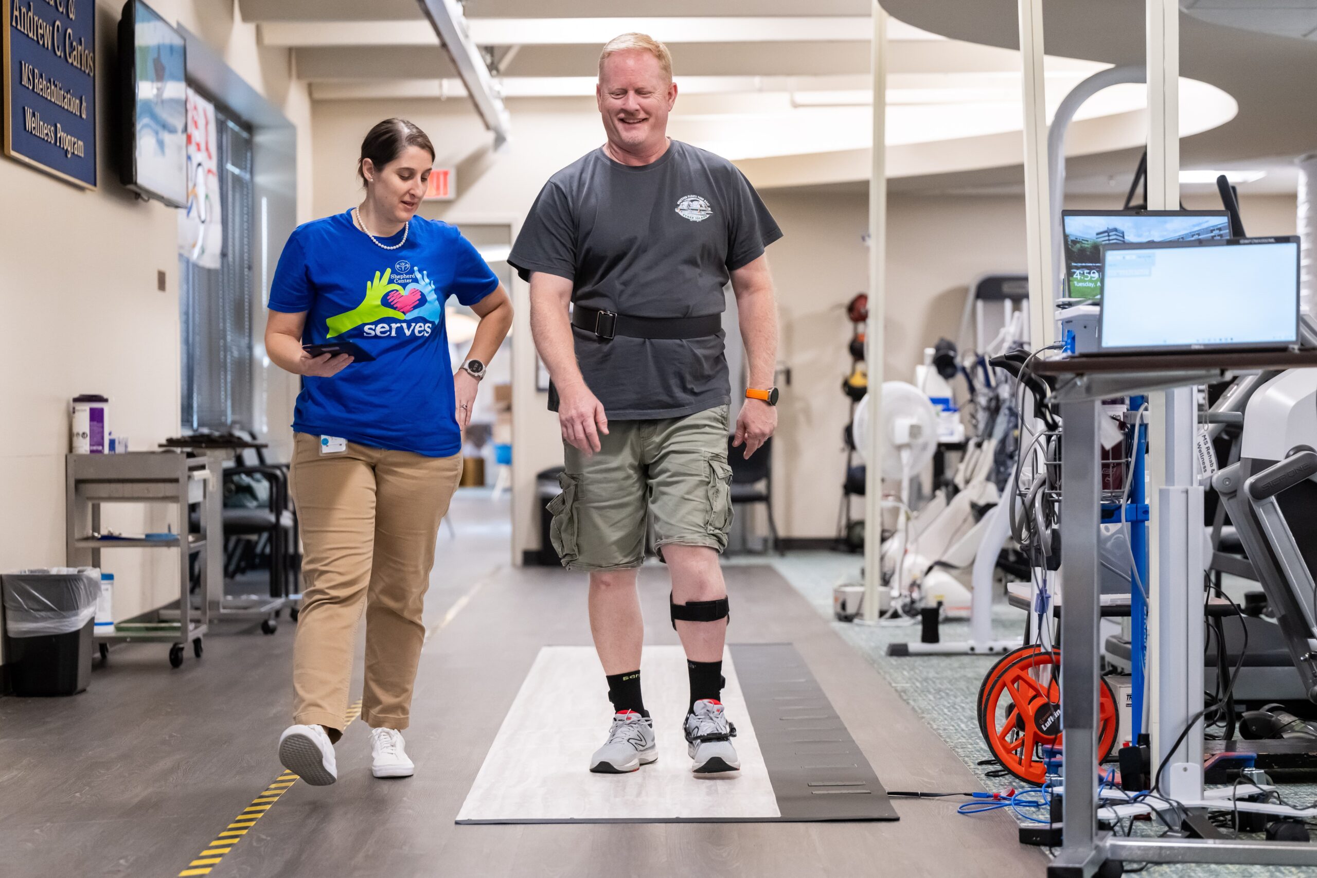 A man with a knee brace walks in a rehabilitation facility alongside a female physical therapist, who is offering guidance. Exercise equipment and a laptop are visible in the background.