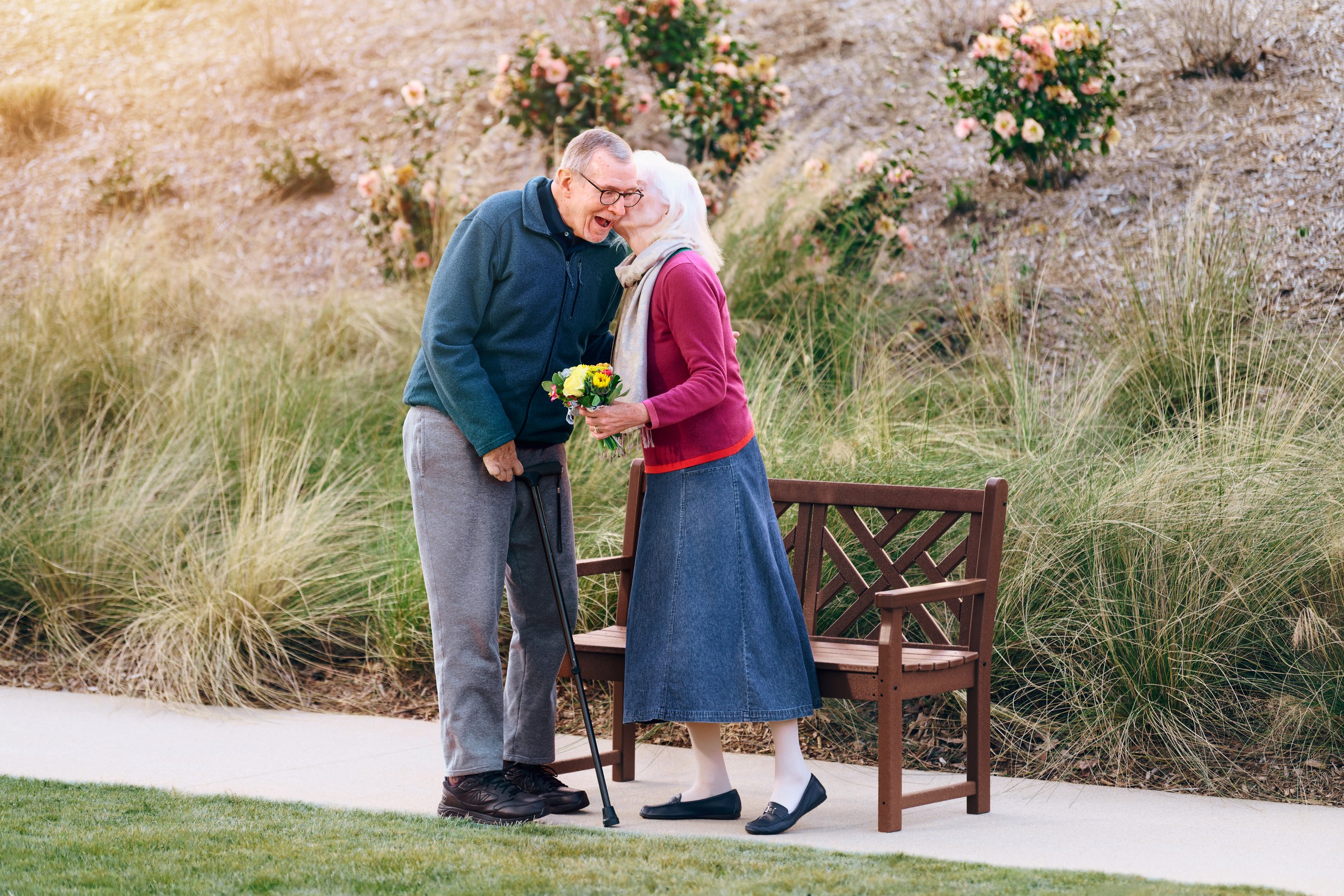 A couple in their 70s stands smiling by a wooden bench outdoors. The woman, holding yellow flowers, leans in to whisper to the man, who uses a cane. They are surrounded by grass and blooming rose bushes.