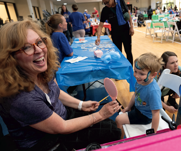 A woman with glasses smiles while holding a pink mirror for a young boy with a painted face, who is sitting on a chair at a lively indoor event with crafts and tables in the background.