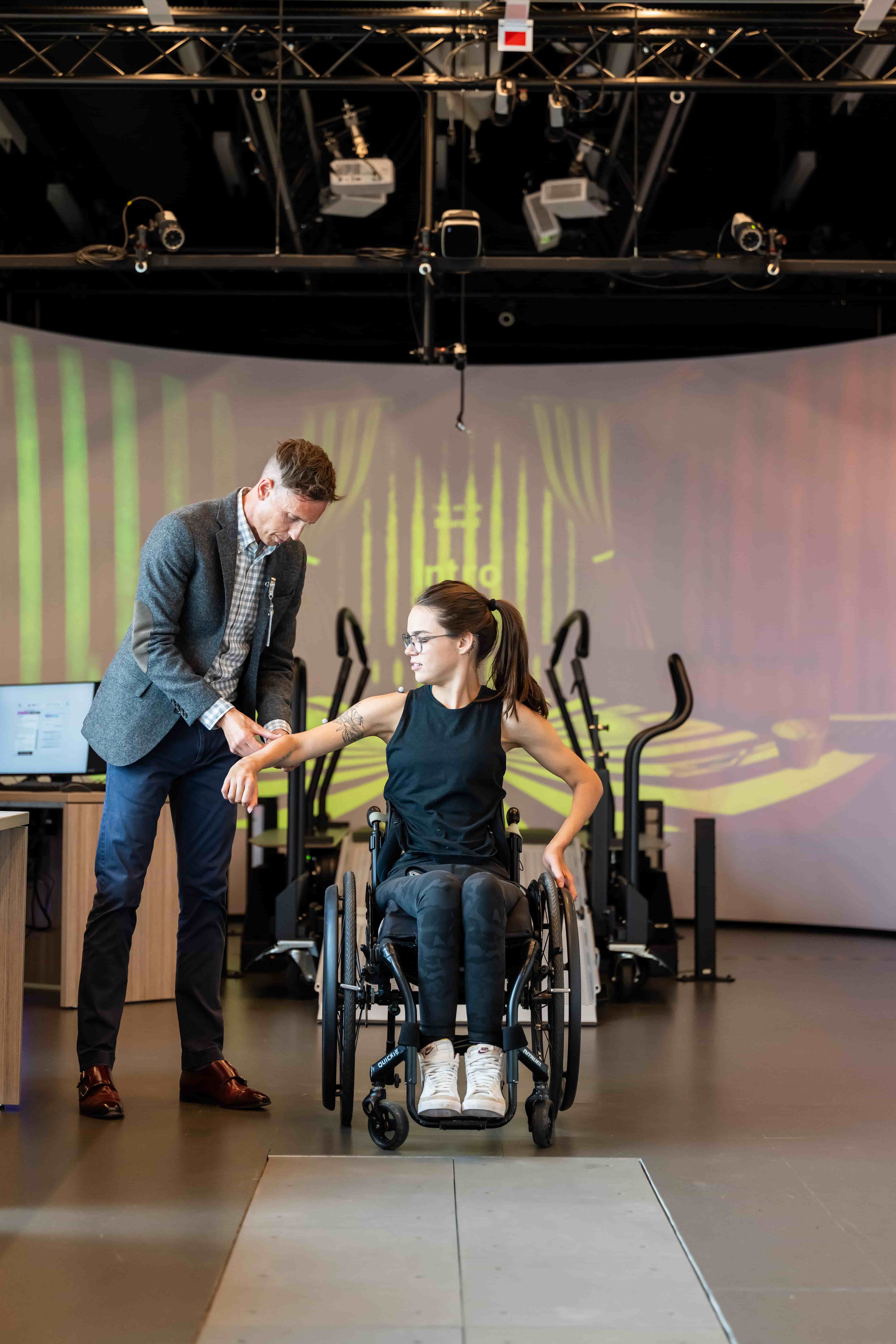 A man assists a woman in a wheelchair in a modern, well-lit room with exercise equipment and a patterned projection on the wall behind them. The woman looks focused as the man supports her arm.
