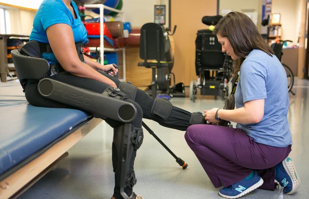 A physical therapist kneels beside a seated person wearing a robotic exoskeleton, adjusting the device on their leg in a rehabilitation center.