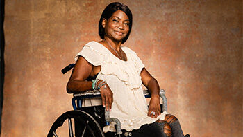 A woman in a wheelchair smiles at the camera. She is wearing a light-colored blouse and black pants, with her hair styled neatly and bracelets on her wrist, sitting against a warm, textured background.