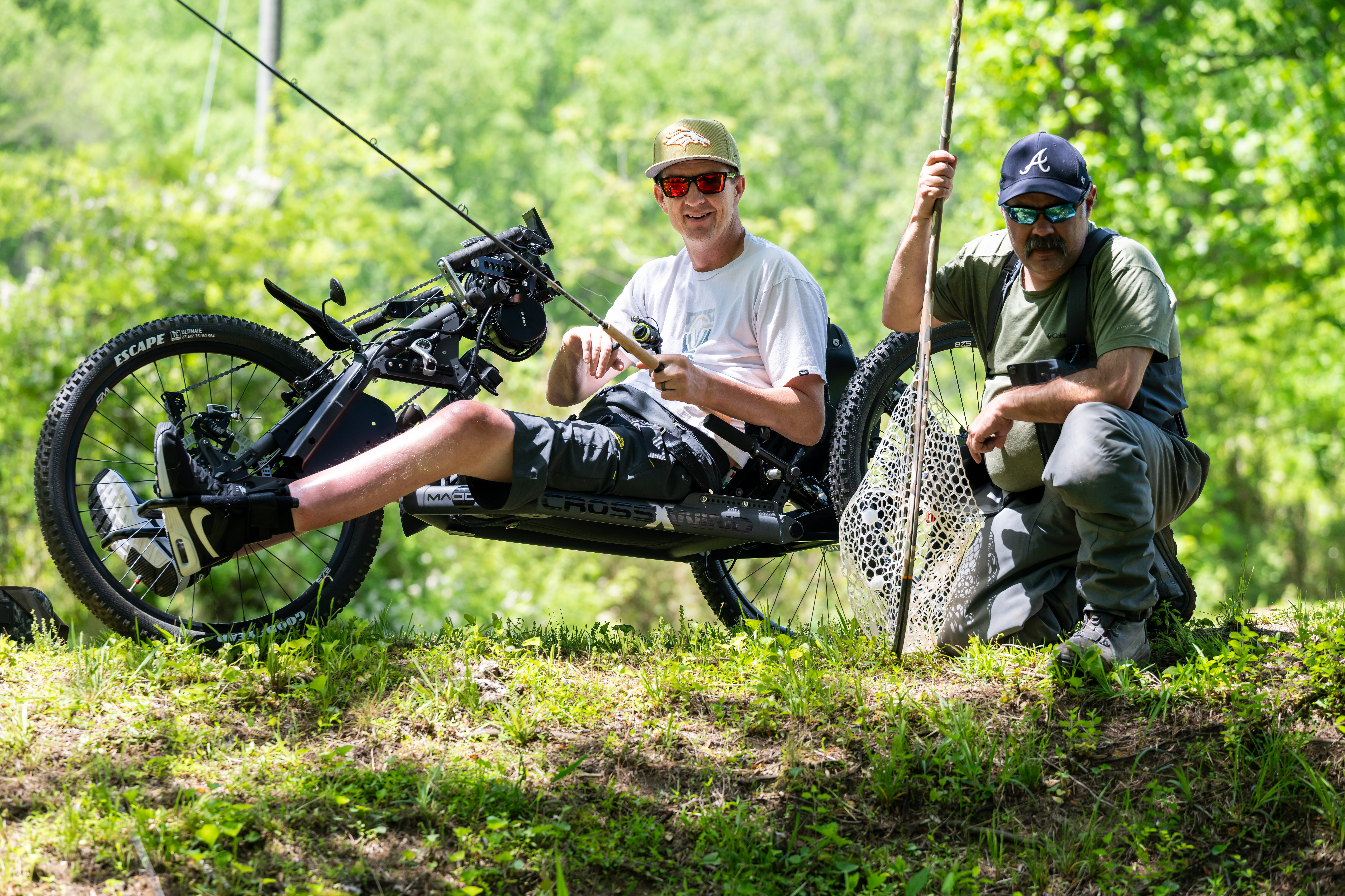 Two men enjoy fishing by a grassy riverbank. One is seated on an adaptive bike, holding a fishing rod. The other is crouching, wearing waders and holding a fishing net. The scene is set amidst lush green trees and sunlight.